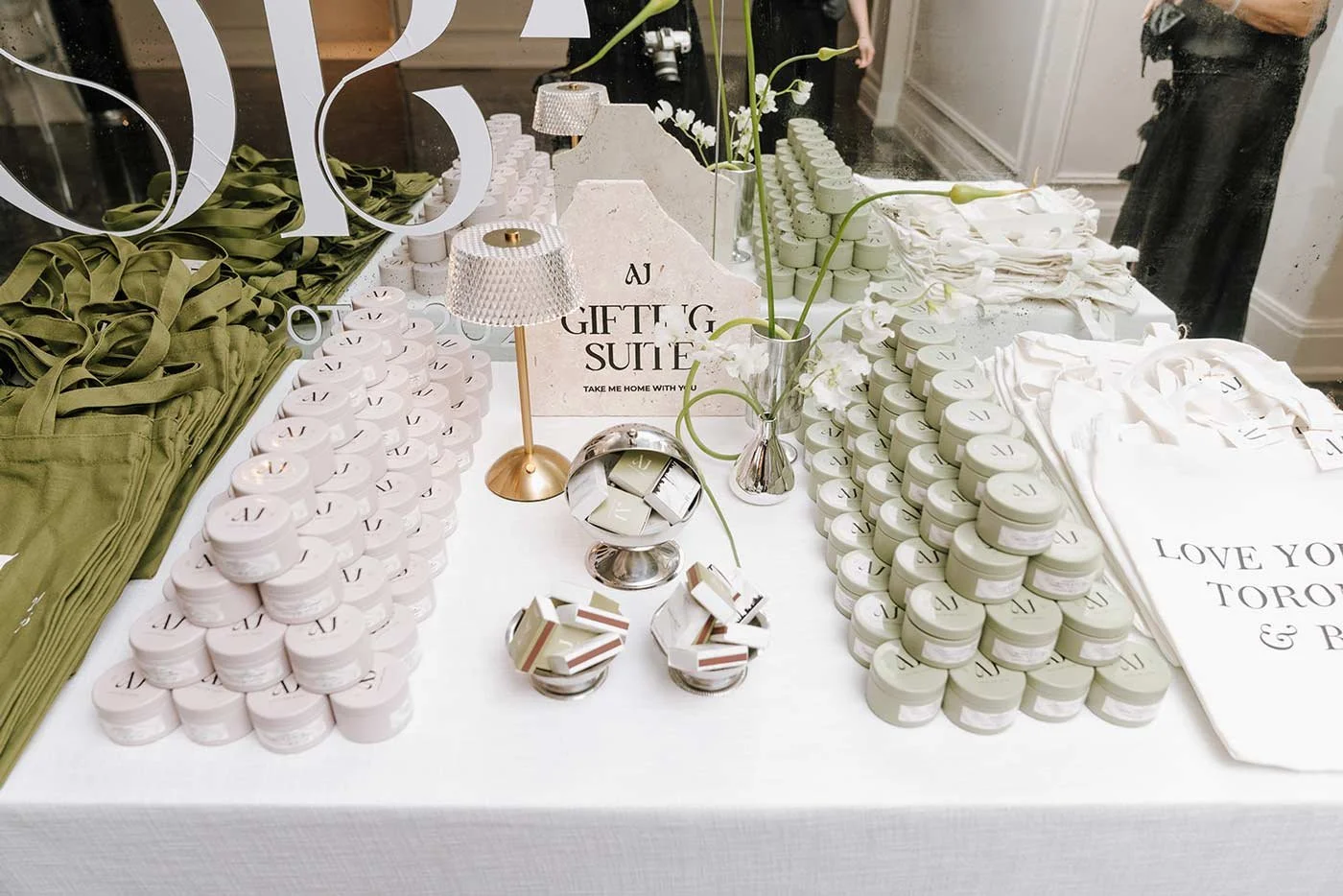 Display table with stacks of small round containers, folded fabric items, flowers in a vase, and sign reading 'AJ Gift & Suit'.