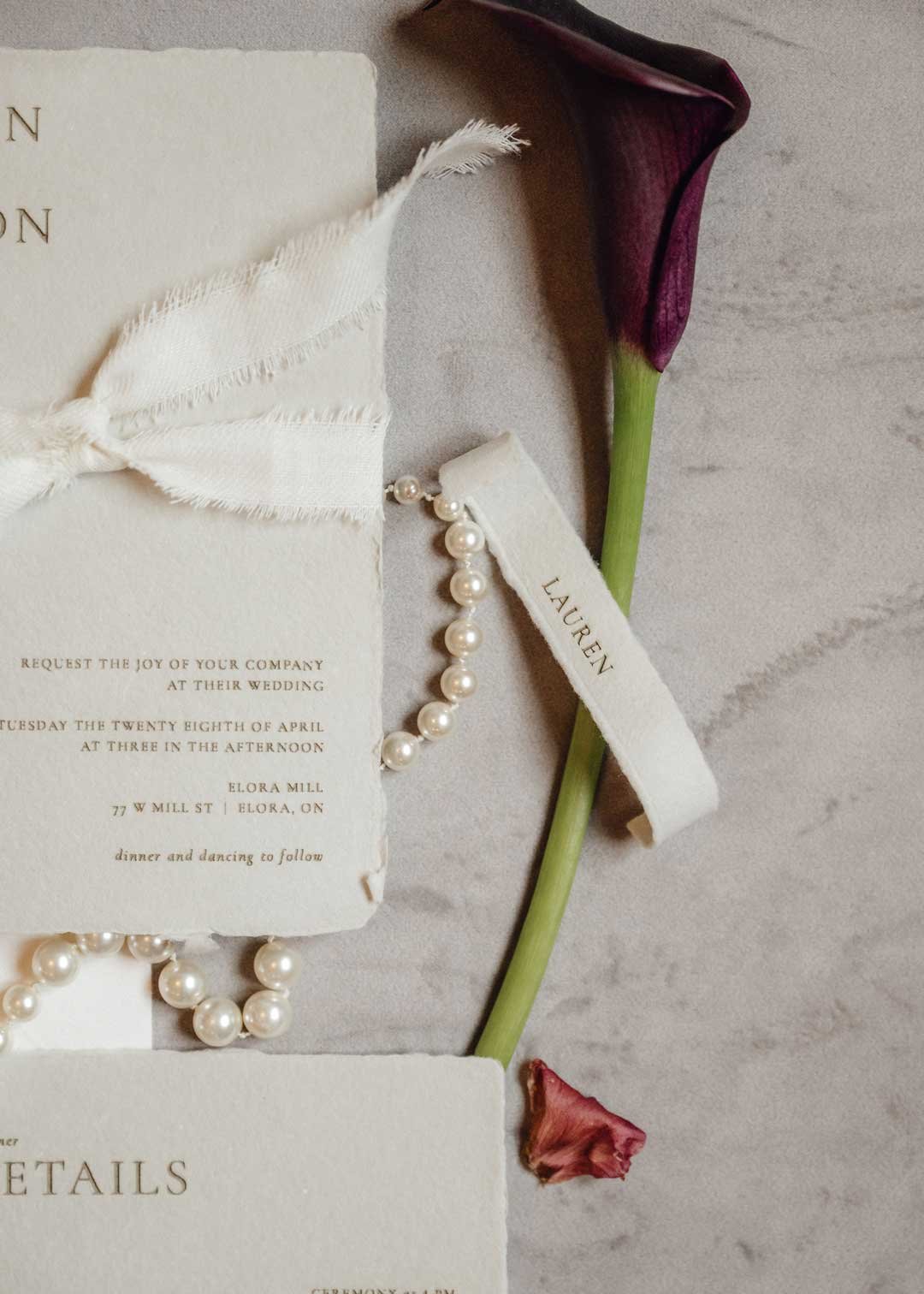 A wedding invitation with a white ribbon, pearl necklace, and a dark purple calla lily on a light gray surface.