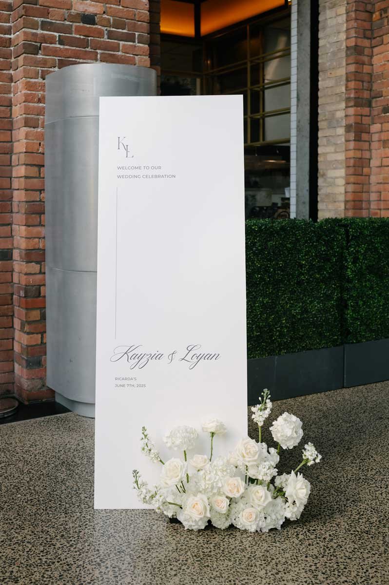 Wedding welcome sign with white flowers at the base, placed outside a brick building.