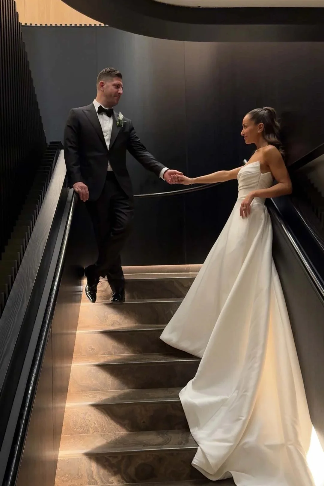 A bride and groom holding hands on an escalator in a modern, dark-colored interior space, with the bride in a white gown and the groom in a black tuxedo.