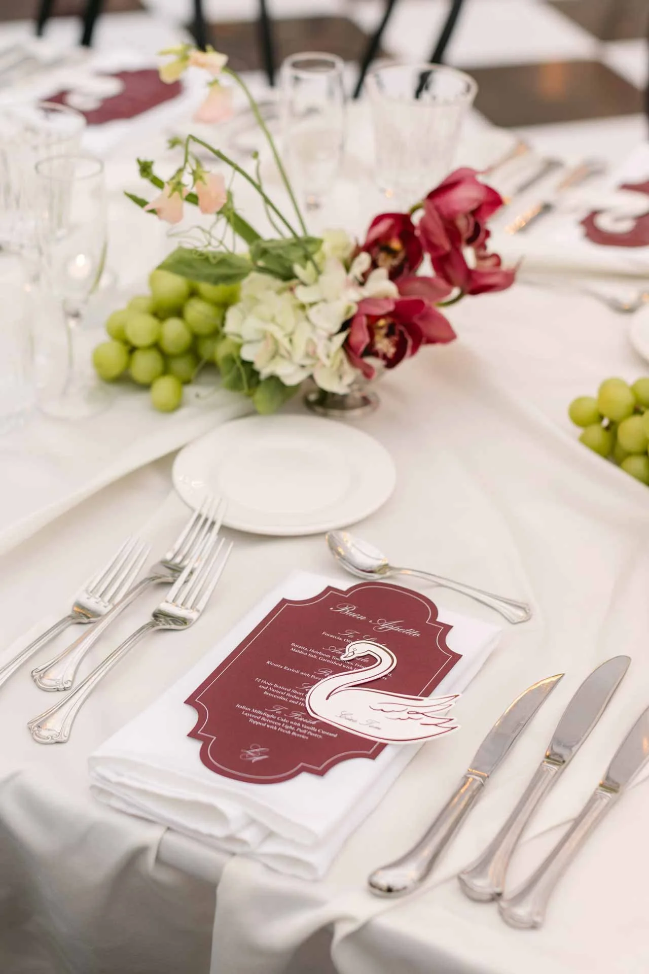 A elegantly set dining table with cutlery, a white napkin, a menu card with a swan graphic, and a floral centerpiece with pink and white flowers, green grapes, and glassware.