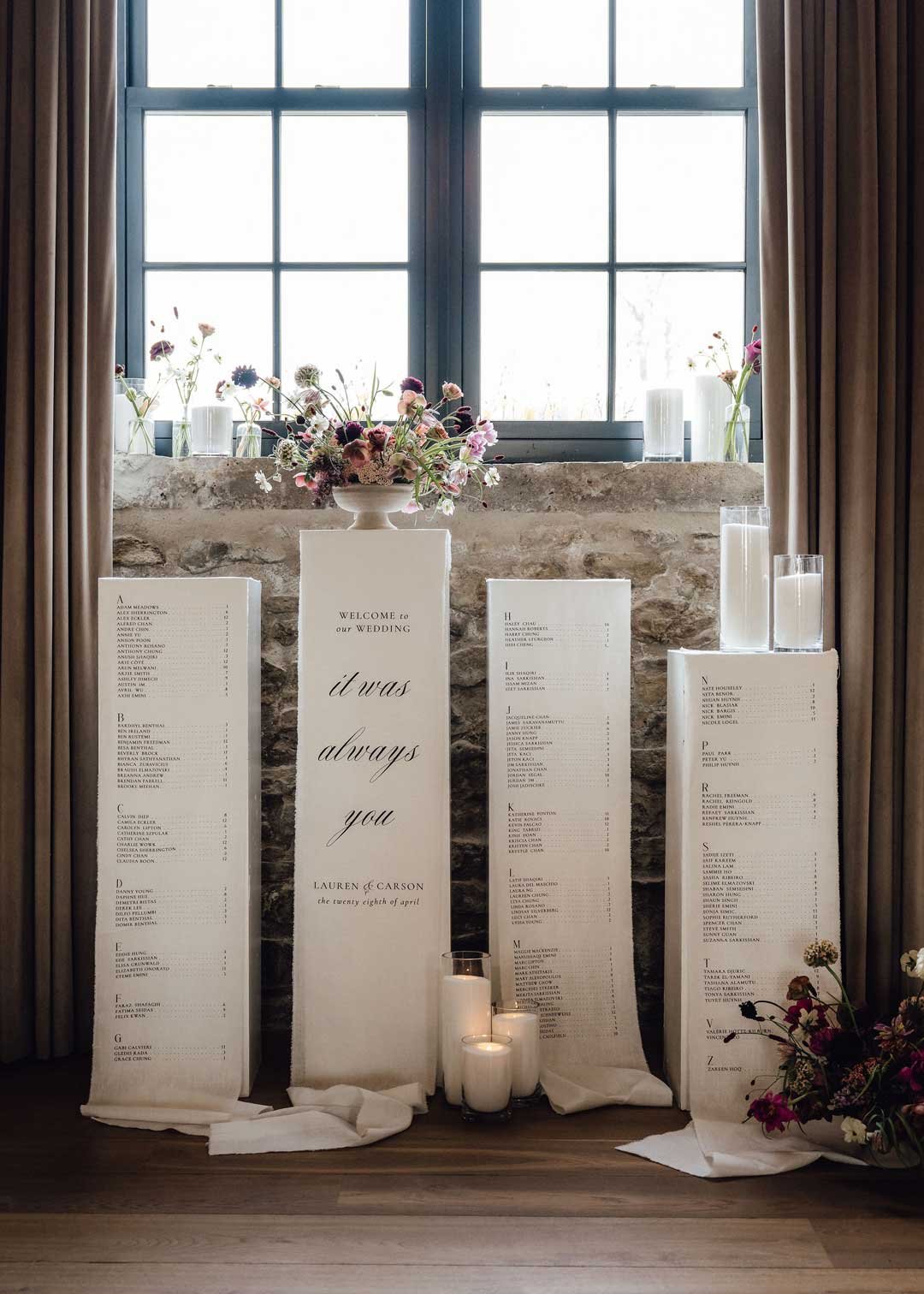 A wedding seating chart display with four white panels, each listing guest names alphabetically, decorated with floral arrangements and candles, against a window with curtains.