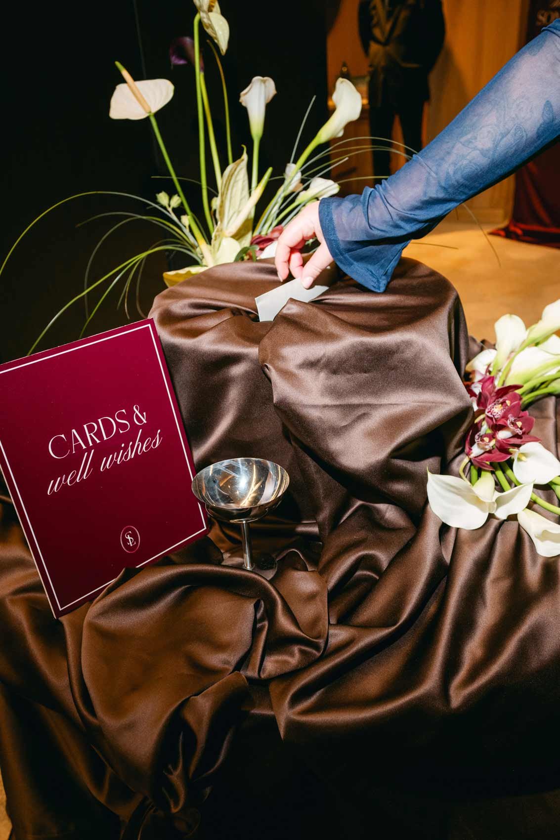 A person in a blue sleeve drops a card into a box at a wedding or celebration. The box is draped with brown fabric and surrounded by white flowers, including orchids. A sign reads "CARDS & well wishes."
