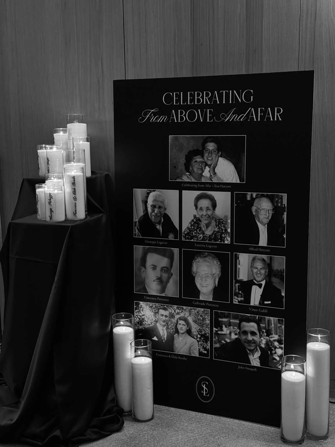 A memorial display board with photographs of multiple people, surrounded by lit white candles on the floor and a table, with a black backdrop and a black and white photo collage, in a solemn setting.