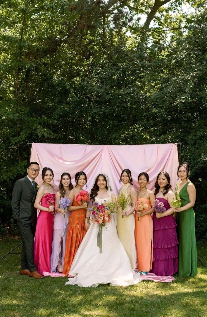 A group of nine people, including a bride in a white dress, standing outdoors in front of a pink backdrop and lush green trees. The group is dressed in colorful formal attire, holding bouquets of flowers.