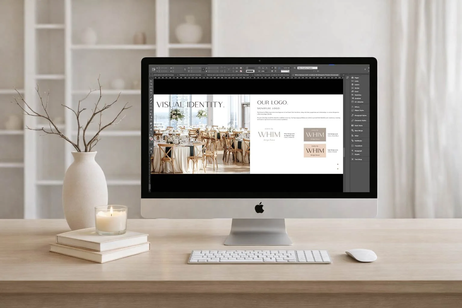A computer monitor on a light wood desk displaying a design project for event branding, with a vase containing branches, stacked books, a lit candle, a keyboard, and a mouse on the desk, and a white bookshelf with decorative items in the background.