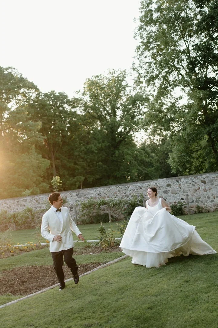 A bride in a white wedding gown and a groom in a white tuxedo walking and smiling on a green lawn with a stone wall and trees in the background during sunset.