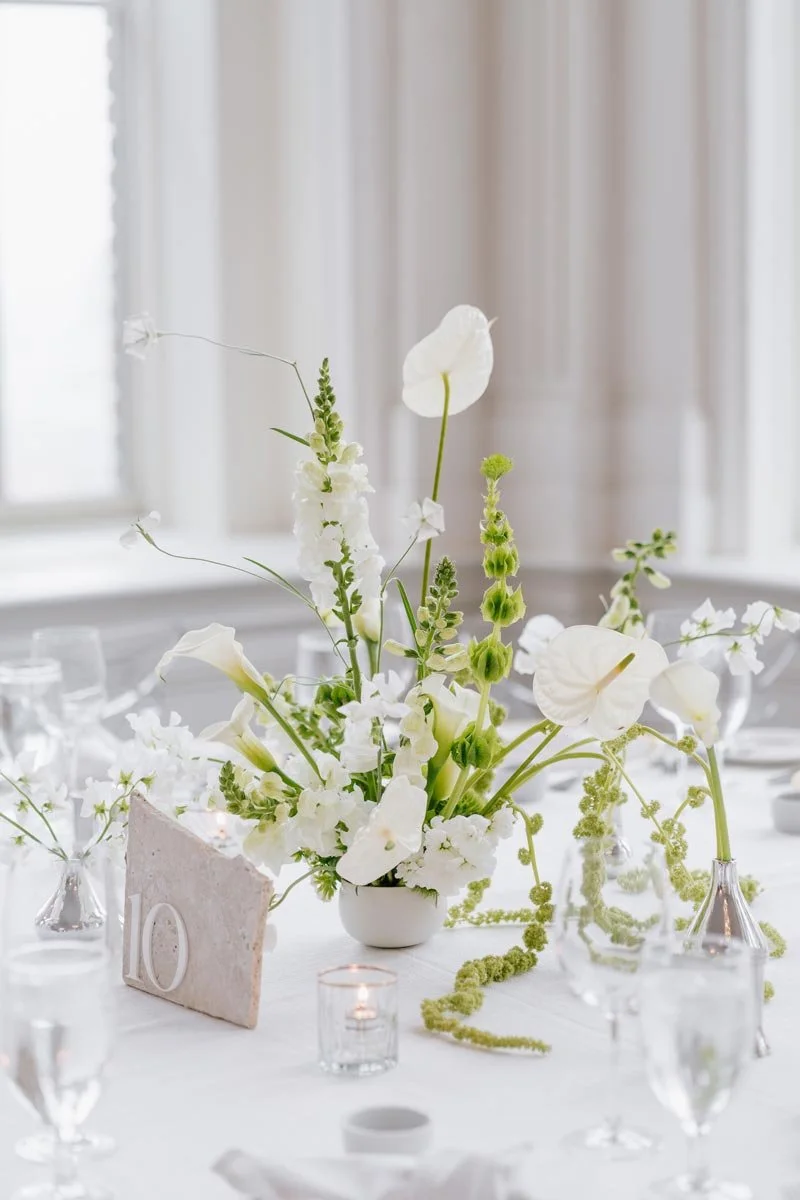 Elegant table centerpiece with white flowers in a small vase, surrounded by candles, glassware, and our stone table number rental on a white tablecloth.