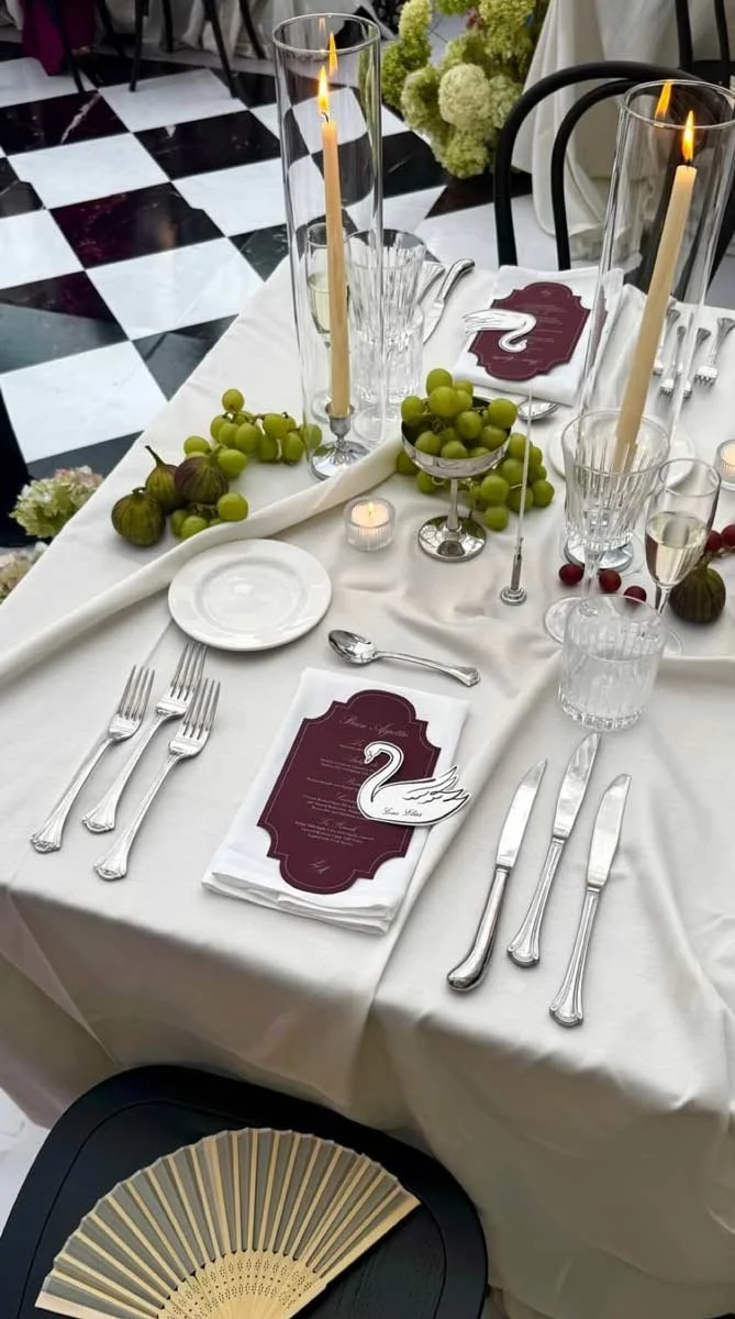 Decorated banquet table set for a formal event with white tablecloth, tall candles in glass holders, plate settings, a menu, grapes, figs, a swan decoration, and glasses for drinks. The background shows a black and white checkered floor.