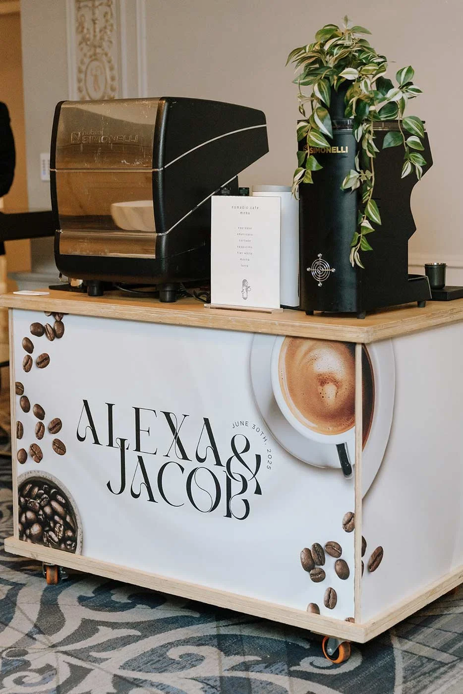 Coffee station with a Simonelli espresso machine, a coffee grinder, a potted plant, and a white menu on a cart that has a design featuring coffee beans and a cup of coffee with the names Alexxa and Jacob, and the date June 30th, 2023.