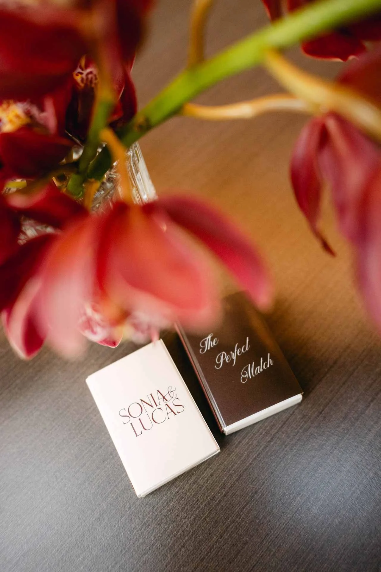 A bouquet of red flowers partially obscures two matchboxes, one labeled 'SONIA & LUCAS' and the other 'The Perfect Match,' placed on a dark surface.