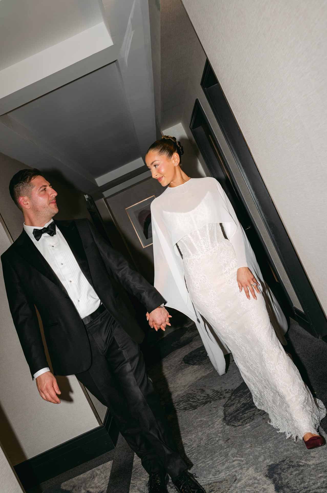 A bride and groom walking together in a hotel hallway, holding hands, dressed in wedding attire. The bride is in a white lace gown with long sleeves, and the groom is in a black tuxedo with a bow tie.