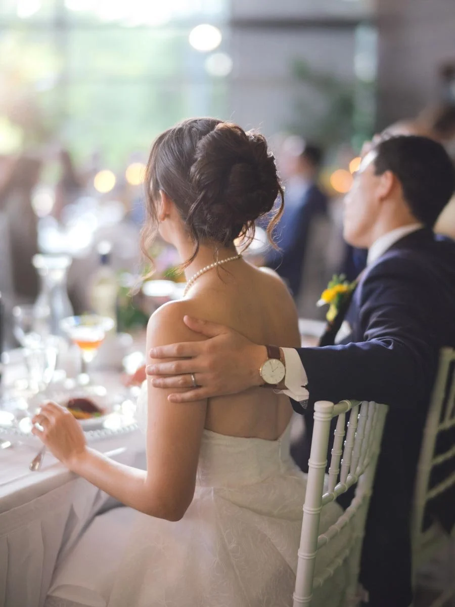 A bride and groom at a wedding reception, sitting close together. The bride has an elegant updo hairstyle and is wearing a strapless wedding dress and pearl necklace. The groom, dressed in a dark suit, has his hand on her shoulder.
