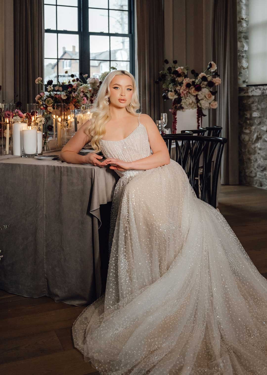 A young woman dressed in an elegant, sparkly wedding gown sitting at a decorated banquet table in a softly lit, rustic decorated venue with large windows and floral arrangements.
