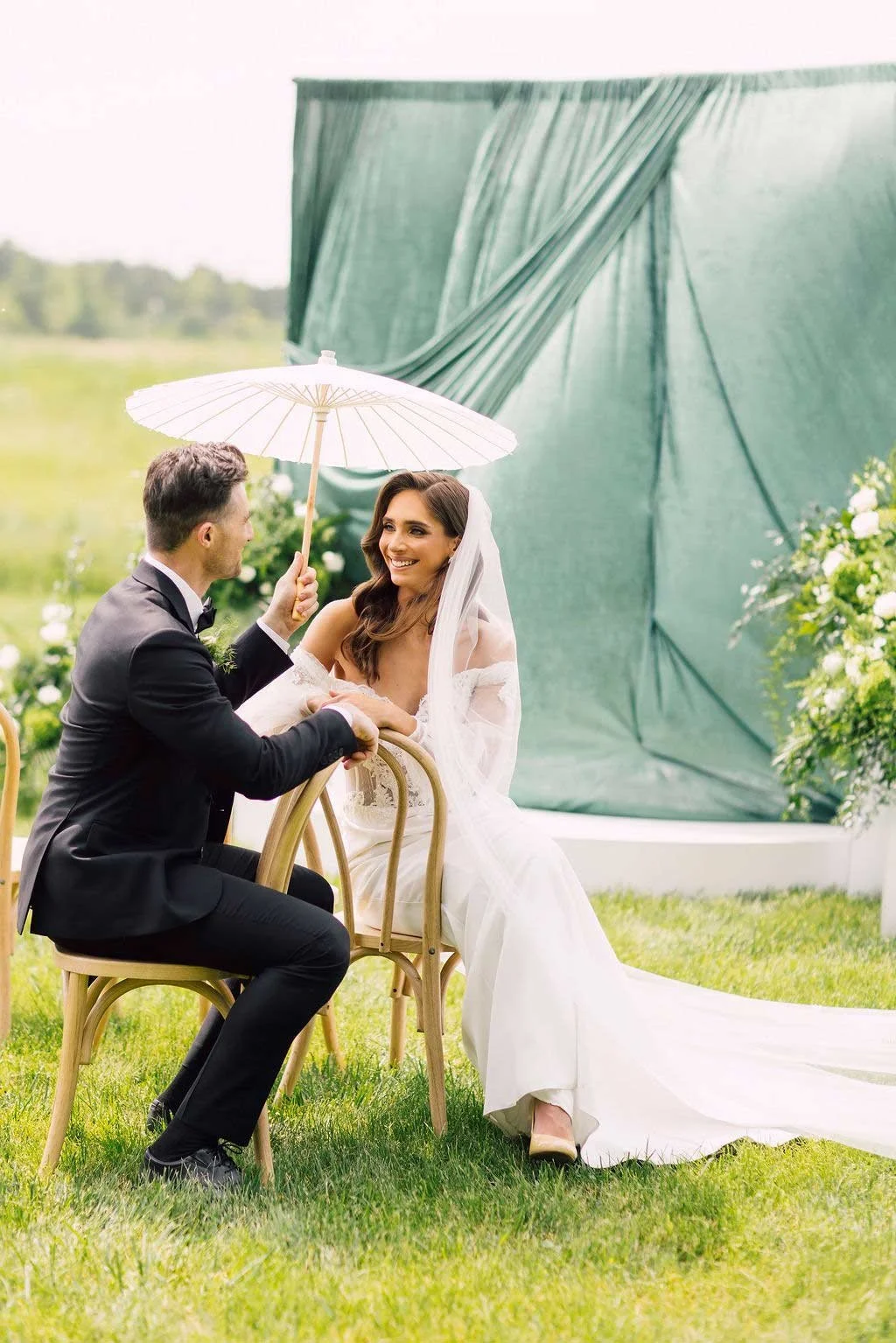 A bride and groom sitting outdoors on chairs, with the groom holding a white umbrella over both of them. The bride is smiling at the groom, dressed in a wedding gown, and the groom is dressed in a black tuxedo. They are surrounded by green grass and decorated with flowers.