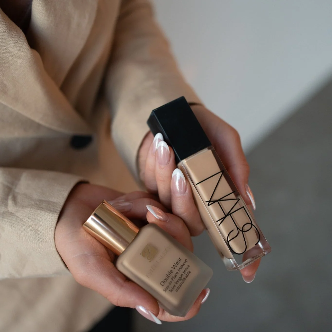 Two hands holding bottles of makeup foundation, one from Estée Lauder and the other NARS. The person is wearing a beige coat with black buttons.