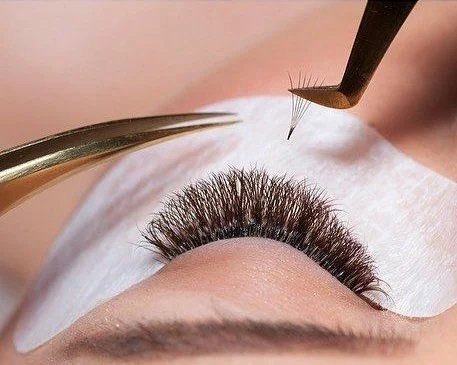 Close-up of an eyelash extension procedure, with a technician attaching synthetic lashes to a person's natural lashes using tweezers and a fine tool.