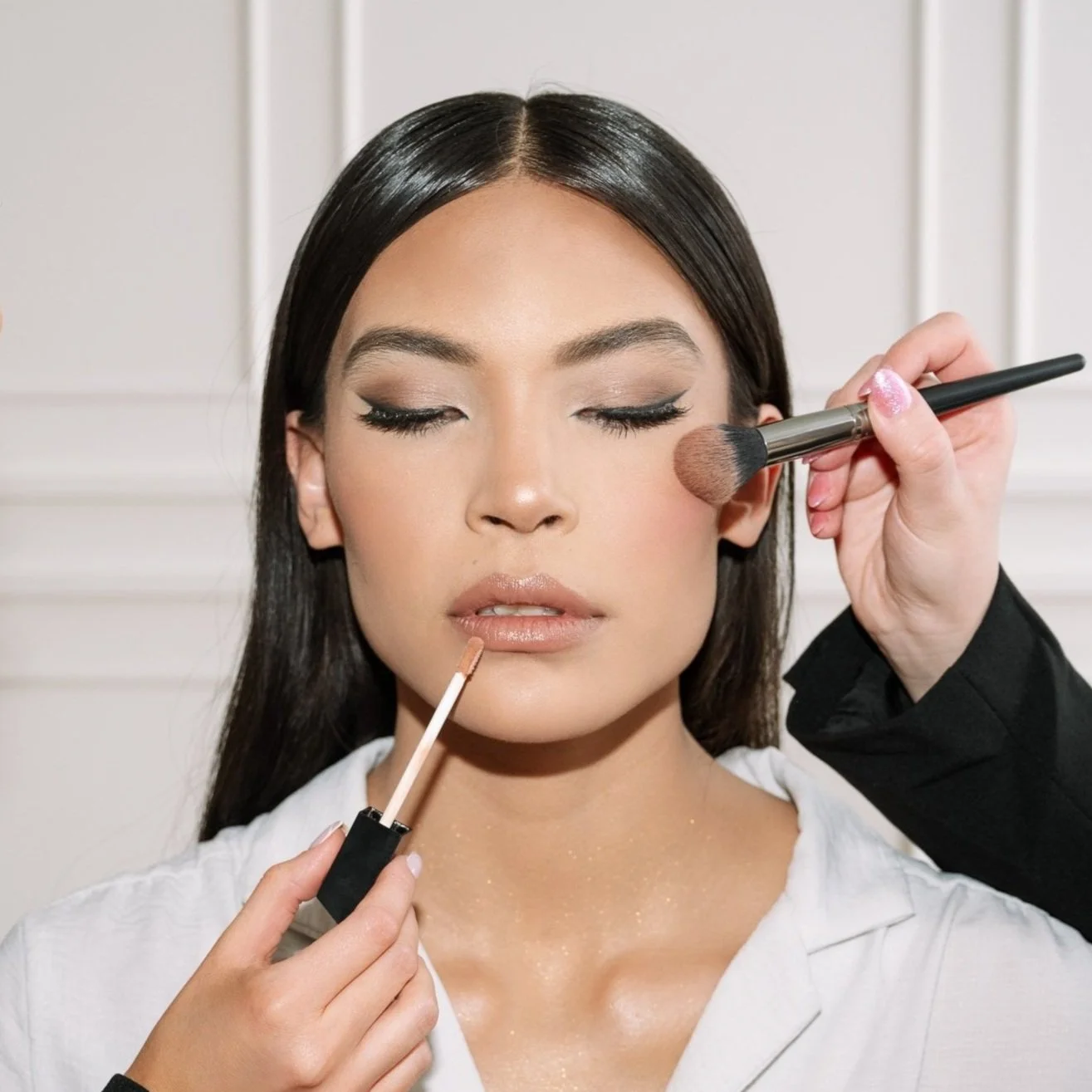 Woman with closed eyes having makeup applied by two people, one applying blush with a brush to her cheek and the other applying lip gloss with a wand.