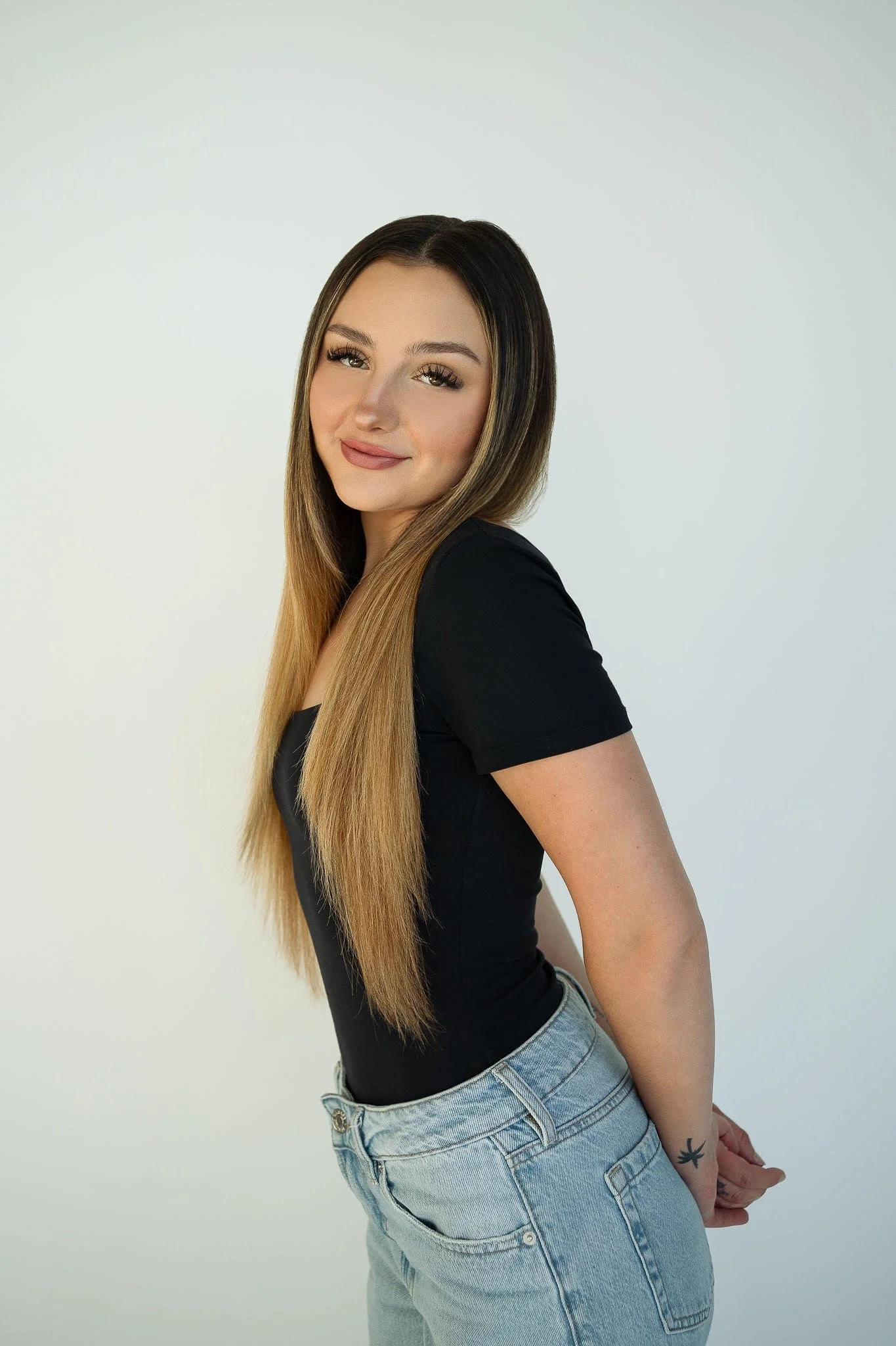 Girl with long brown hair, wearing a black t-shirt and light blue jeans, standing against a plain white background, smiling at the camera.