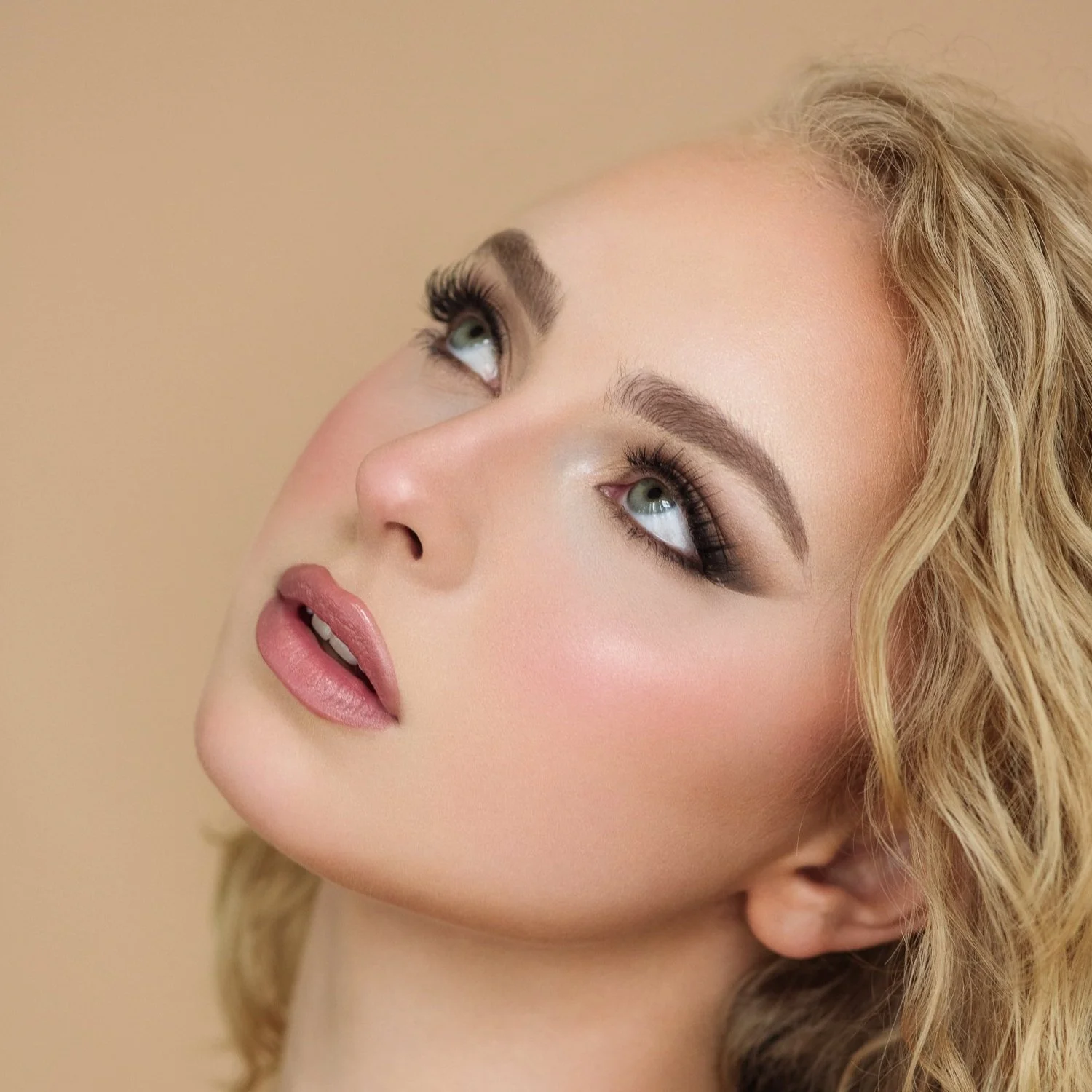 Close-up of a woman with blonde curly hair, glowing skin, and makeup, looking upwards.
