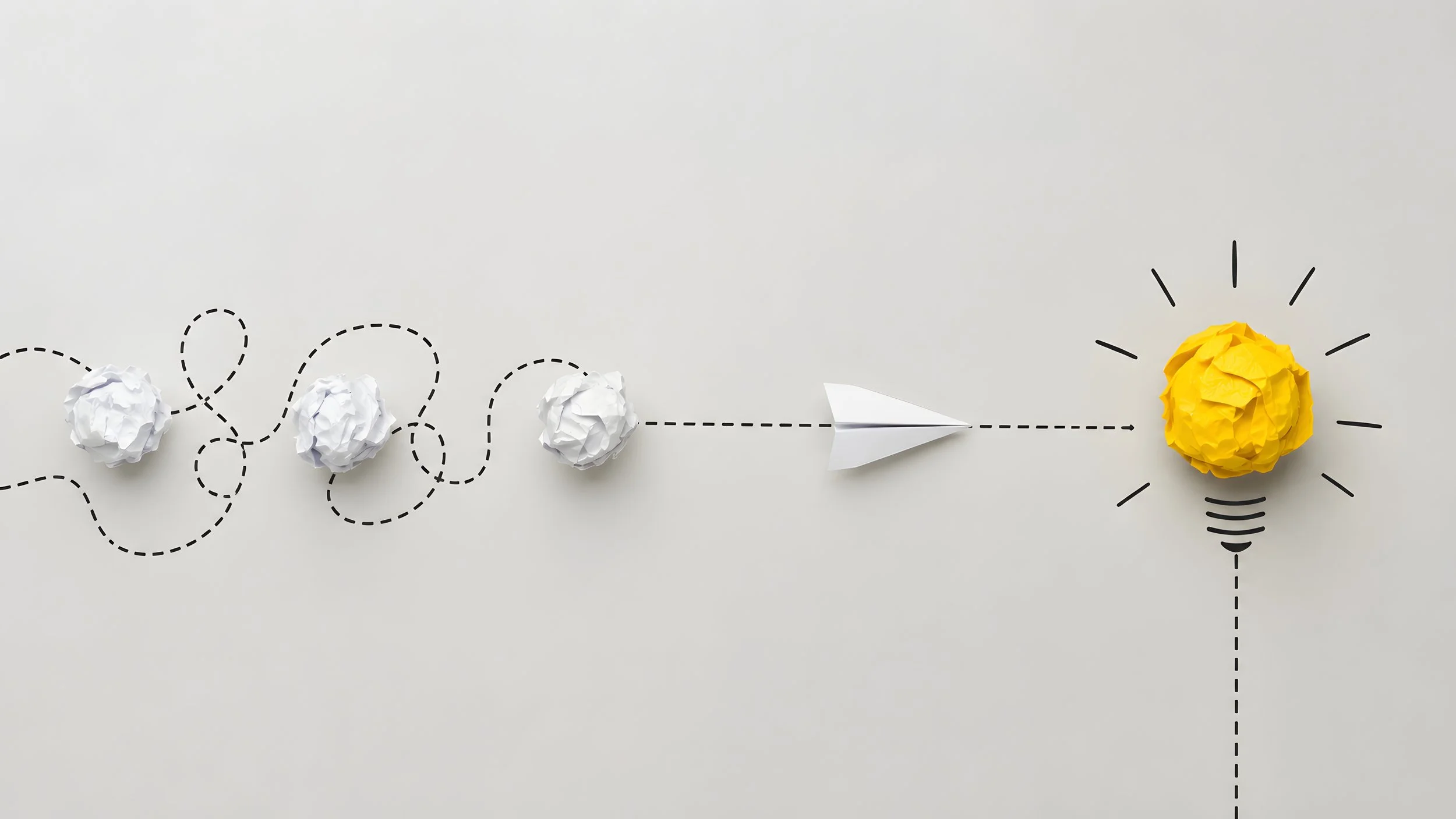 A white background with a dashed black line trail of crumpled white paper balls leading to a yellow crumpled paper ball. The paper ball is designed to resemble a light bulb. Represents the concept of taking a thought into a concept.