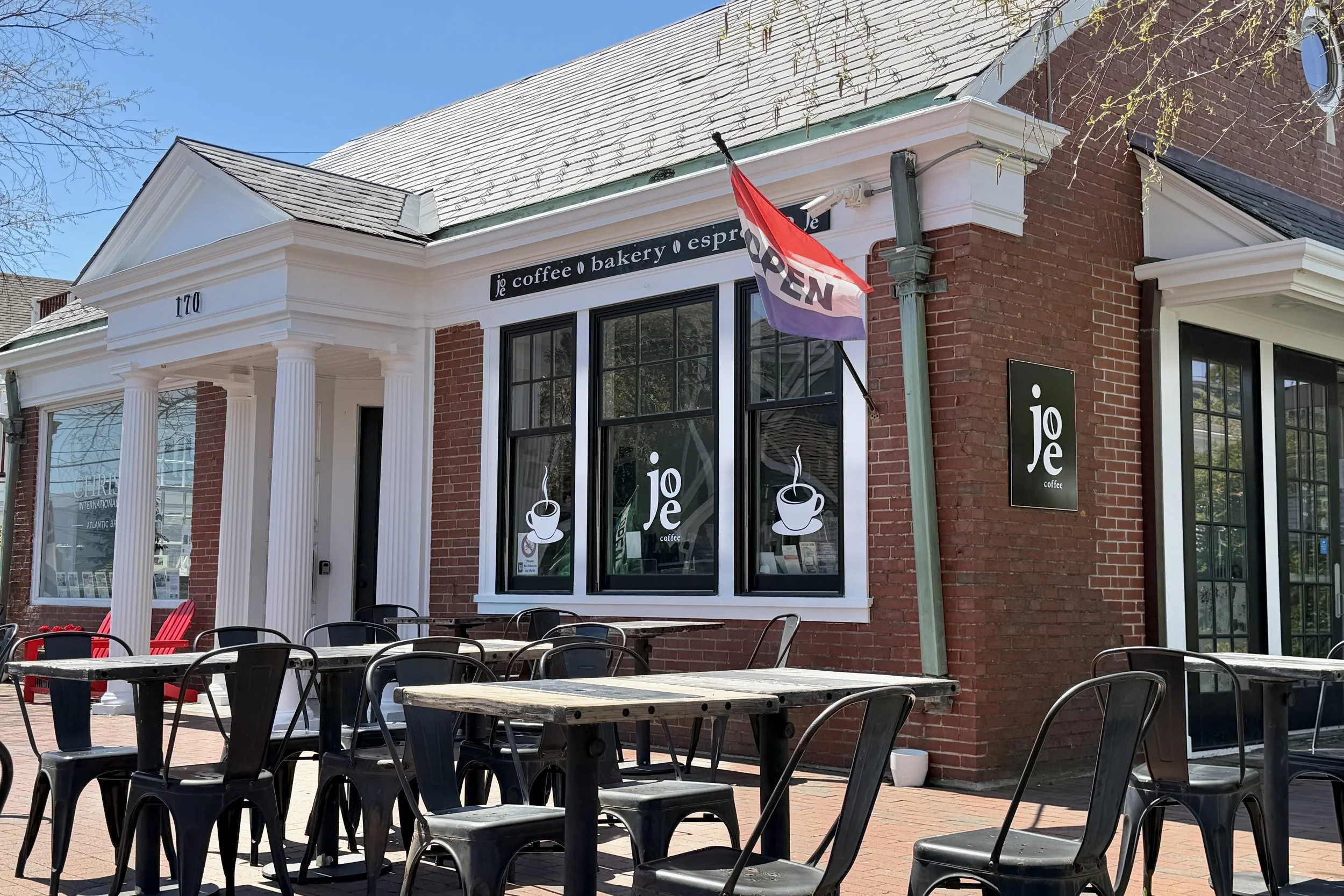The exterior of a coffee shop with outdoor seating, red brick walls, large windows, and a sign that reads 'joe coffee'. There is an 'OPEN' flag, some tables and chairs, and a blue sky overhead.