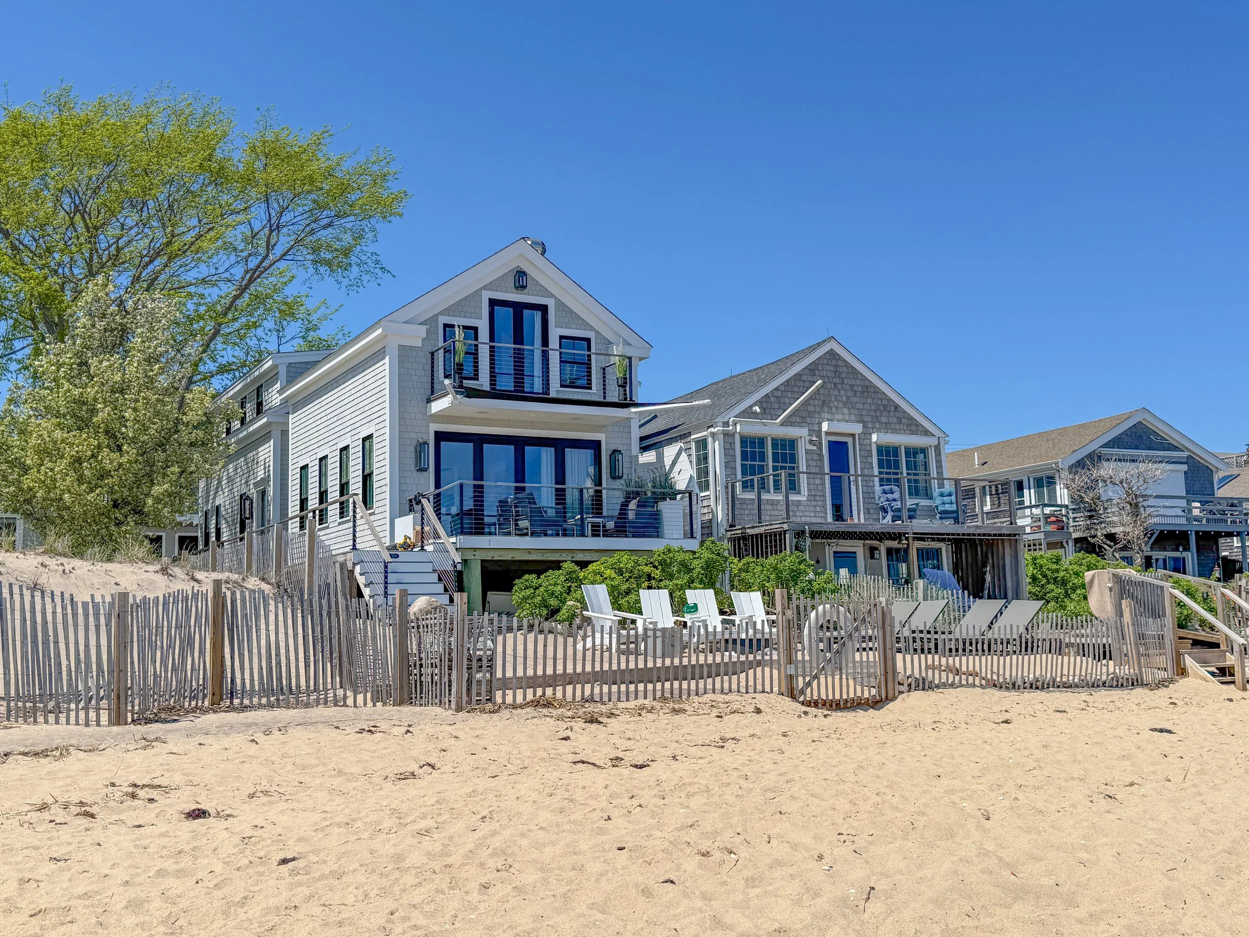 Beach with sand in the foreground and houses with white siding and balconies in the background, trees with budding green leaves, and a clear blue sky.