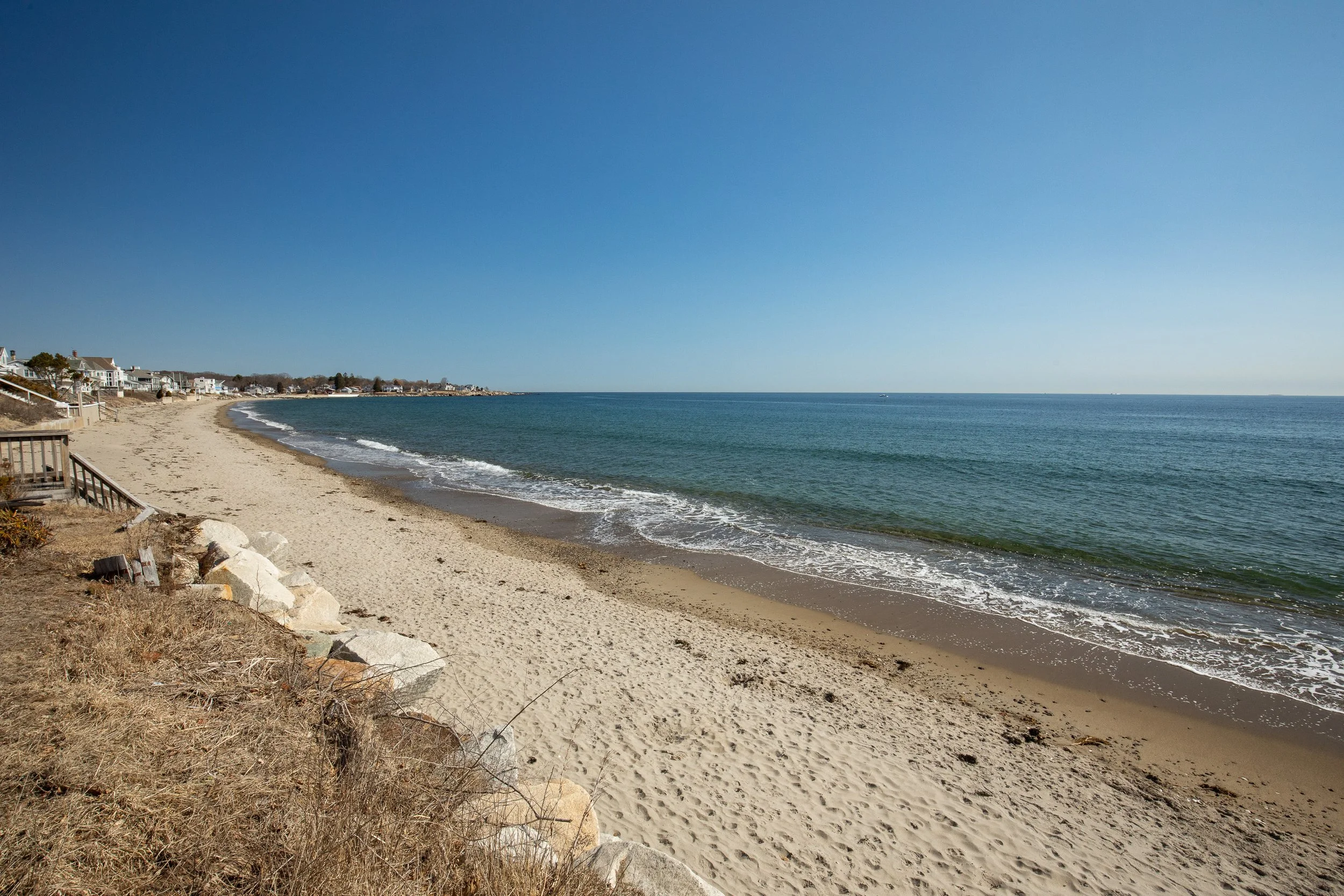 A sandy beach with calm ocean waves, residential houses along the coastline, clear blue sky, and some rocks and vegetation in the foreground.