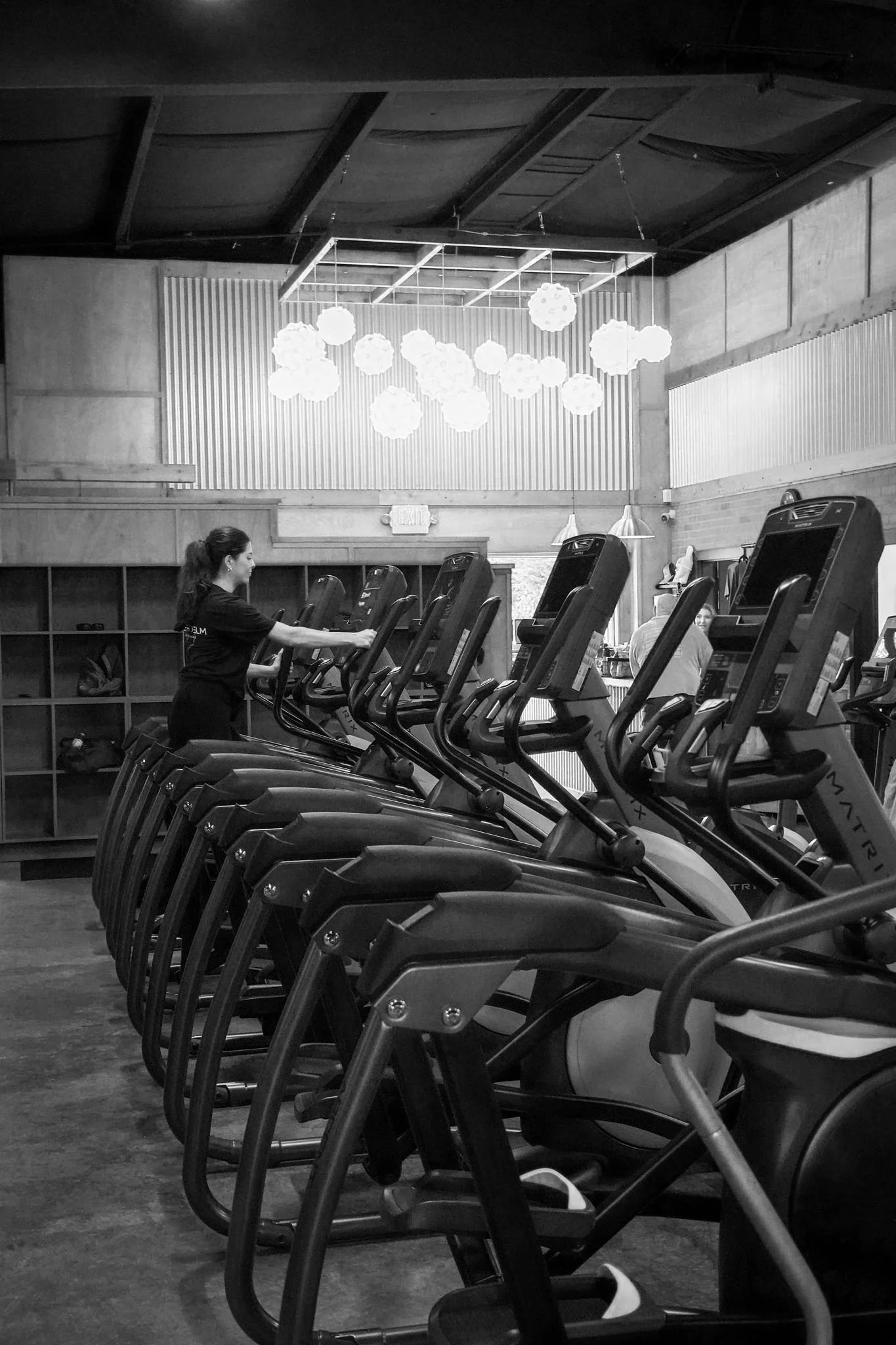 Interior of a gym with a row of exercise machines, including treadmills, in black and white. A woman is using one of the treadmills, and pendant lights hang from the ceiling. There are shelves and a door in the background.