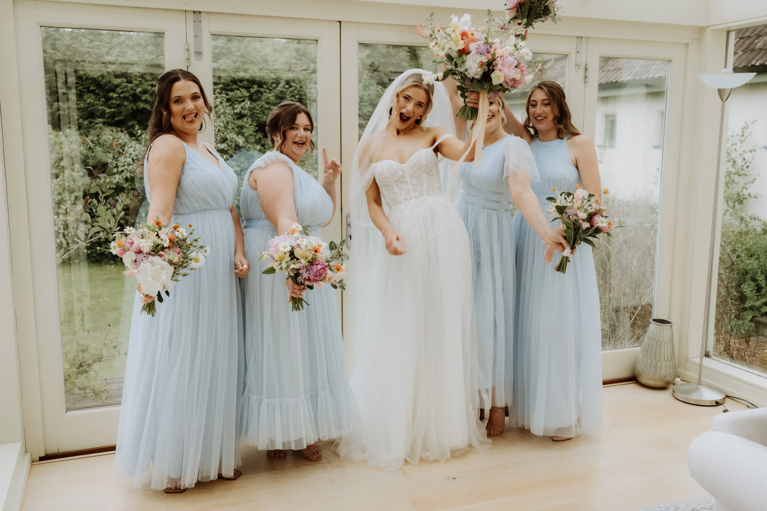 a bride in a white wedding dress standing between four bridesmaids in pale blue bridesmaid dresses waving their wedding bouquets in the air