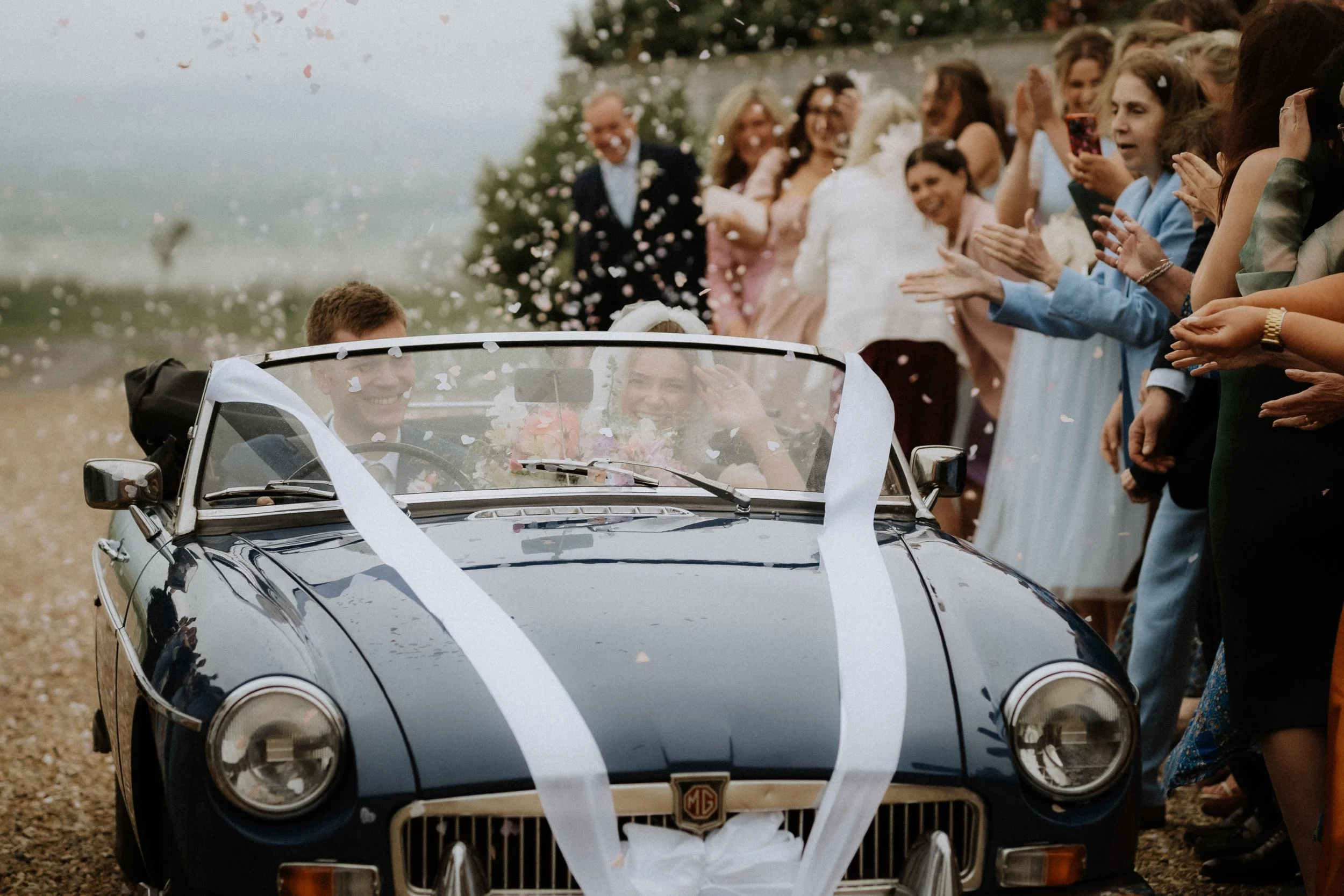 A vintage wedding car driven by a waving bride and groom with confetti being thrown in the air by guests