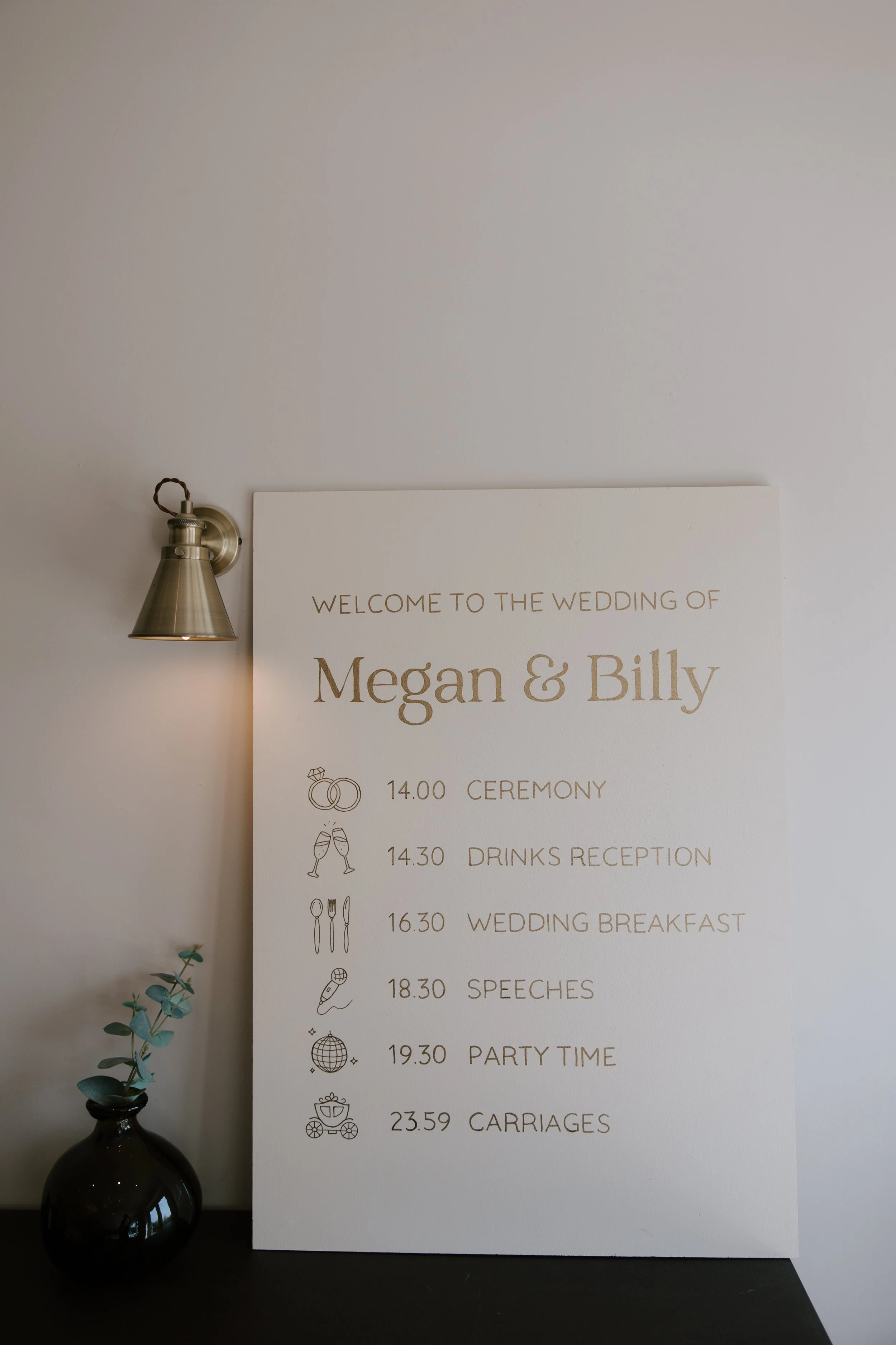 Hand painted wedding order of the day sign with the names Megan and Billy, listing ceremony, drinks reception, wedding breakfast, speeches, party time, and carriages, displayed next to a black vase with green foliage and a wall-mounted lamp.