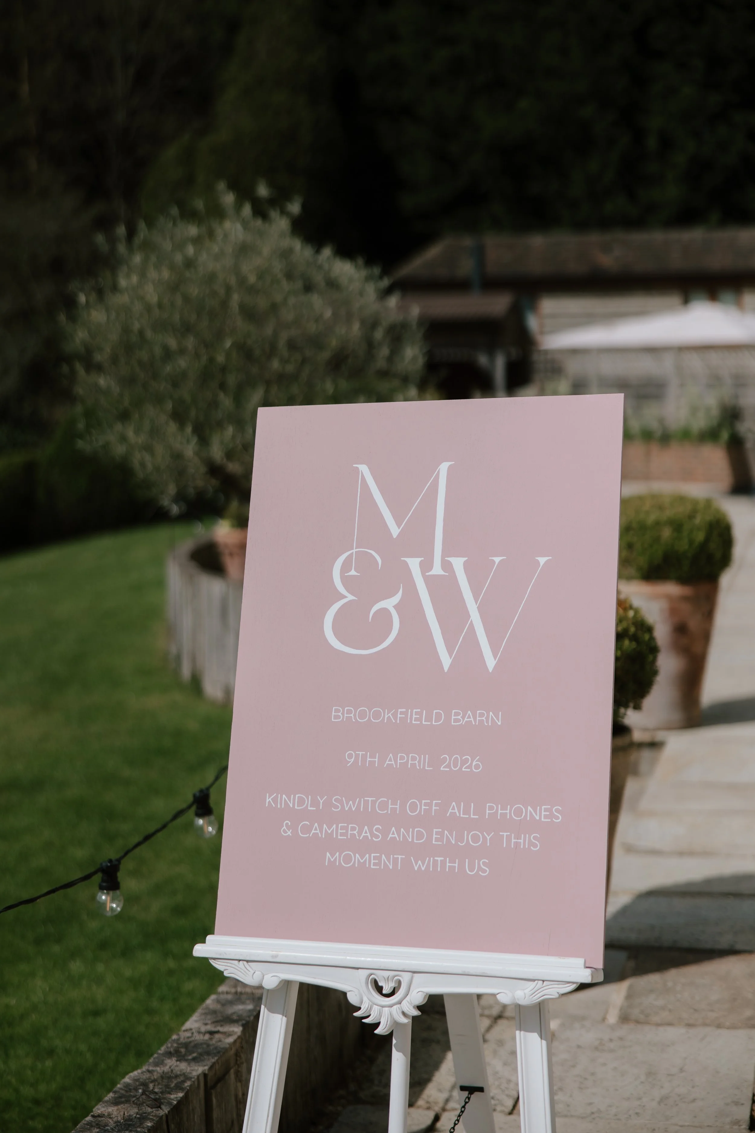 A hand painted pink sign on a white easel at an outdoor wedding with white text reading "Brookfield Barn, 9th April 2026, kindly switch off all phones & cameras and enjoy this moment with us." The background shows a garden, bushes, and buildings.