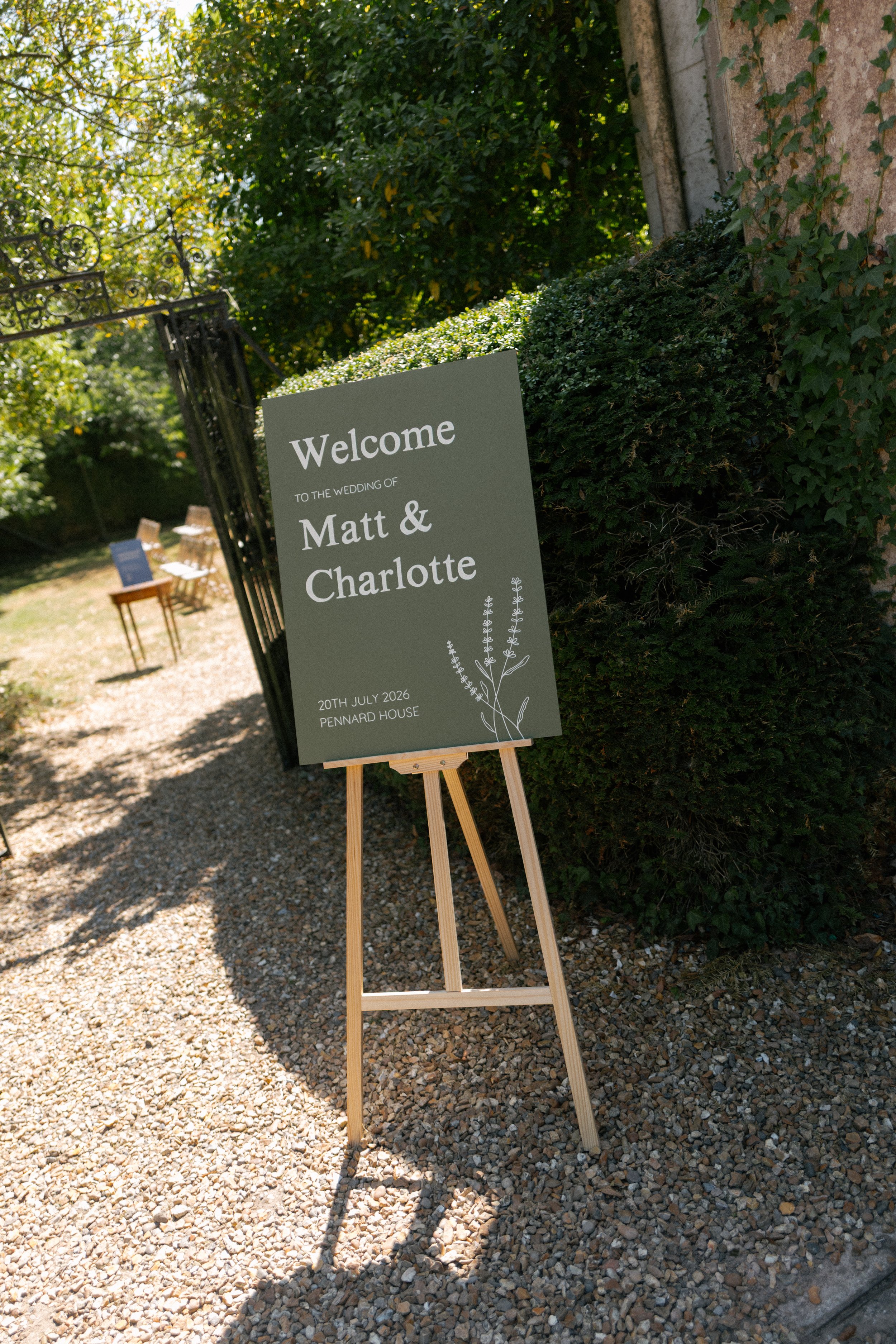 A hand-painted wooden wedding sign with white text. The sign is placed on a wooden easel outdoors for a wedding