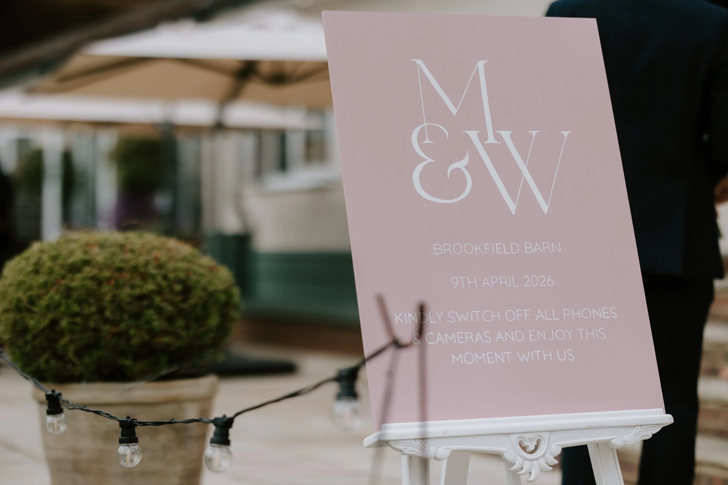 A pink hand-painted wooden wedding sign with white text that reads "M & W, Brookfield Barn, 9th April 2026." The sign is placed on a white decorative stand outdoors, with a grassy area and a building in the background.