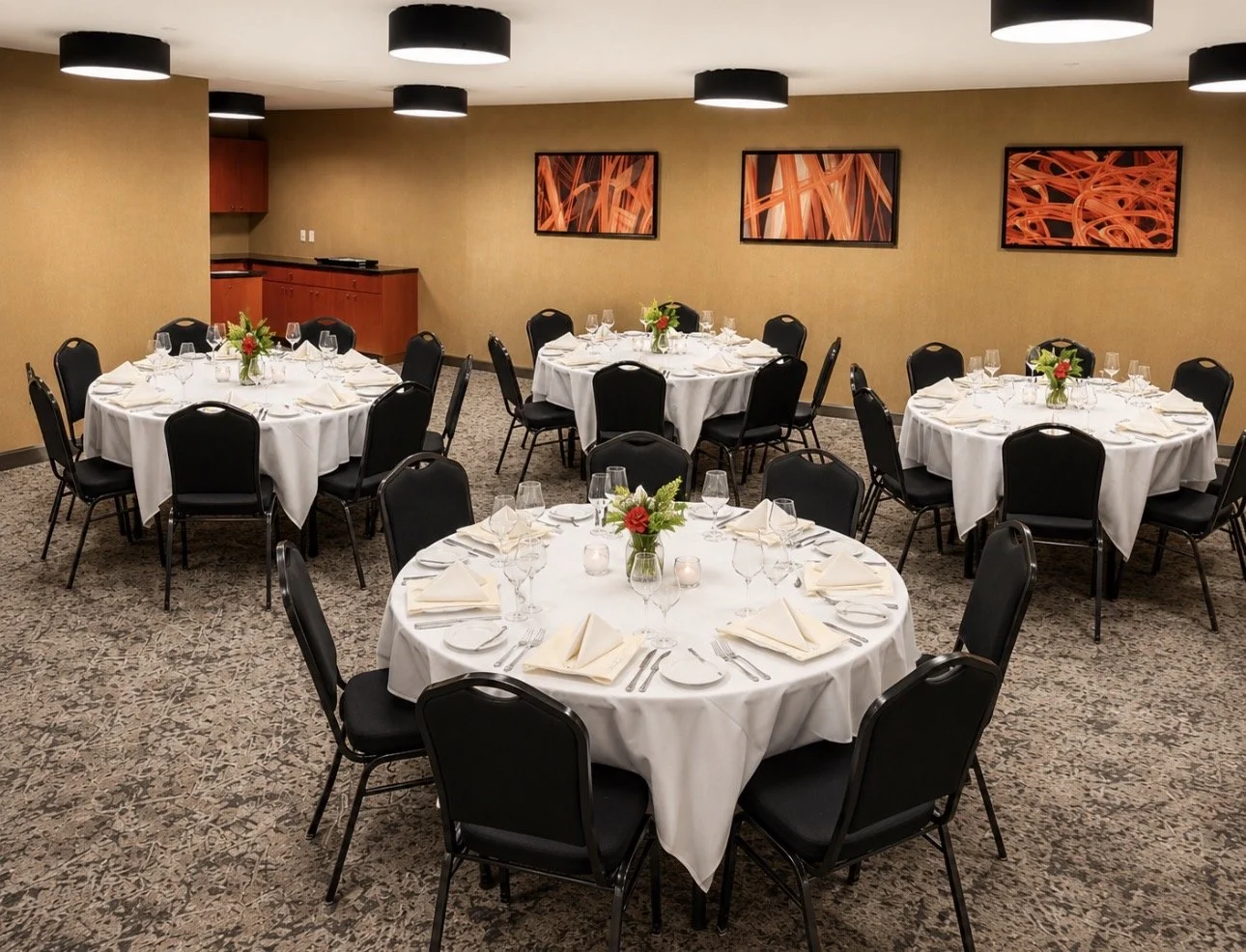 Dining room with round tables covered in white tablecloths, set for a meal with napkins, glasses, and silverware, decorated with small floral centerpieces, surrounded by black chairs, on a carpeted floor, with abstract artwork on beige walls and black ceiling lights.