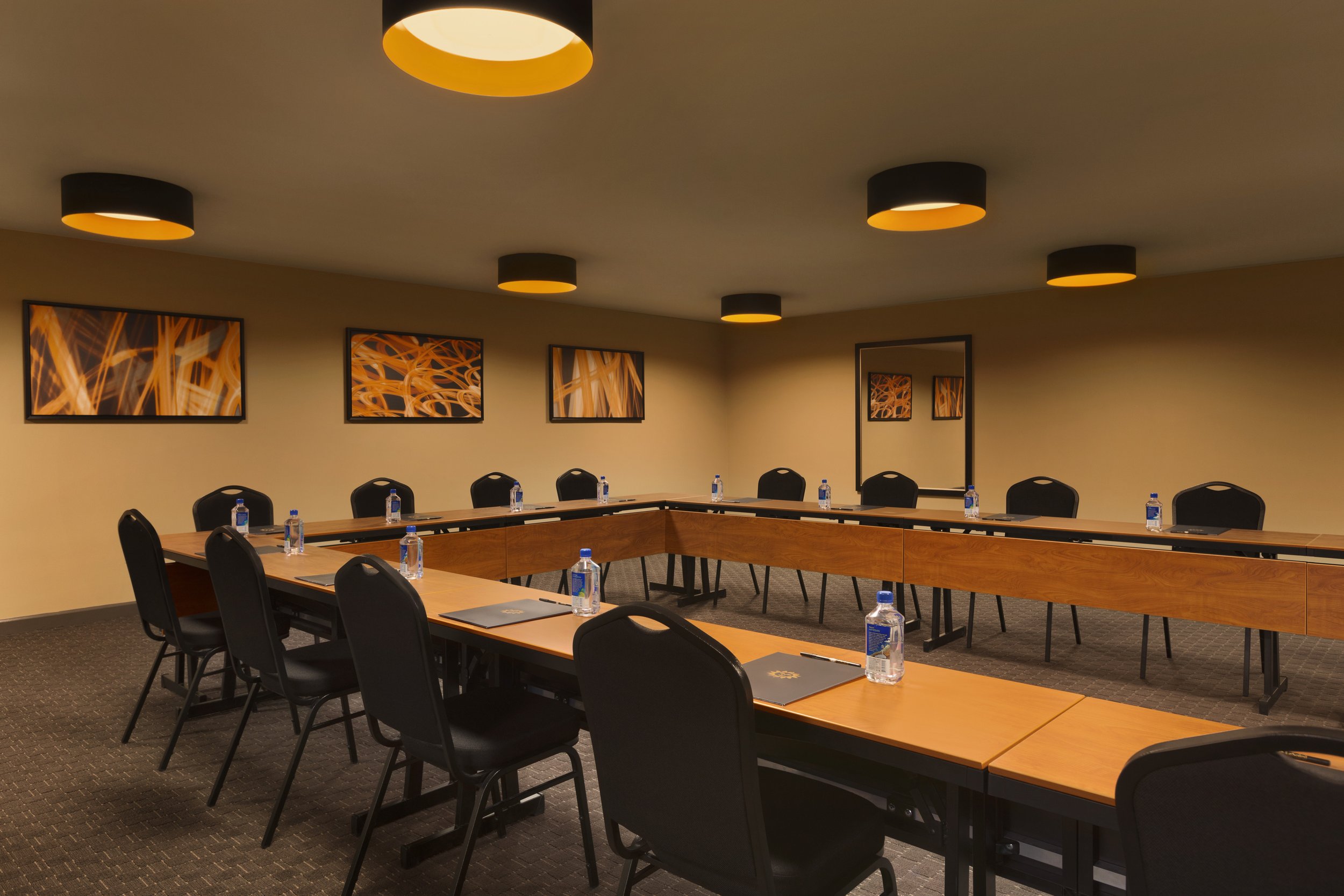 Empty conference room with U-shaped wooden tables, black chairs, bottled water, notebooks, and pens, decorated with orange abstract artwork on beige walls and black ceiling lights.