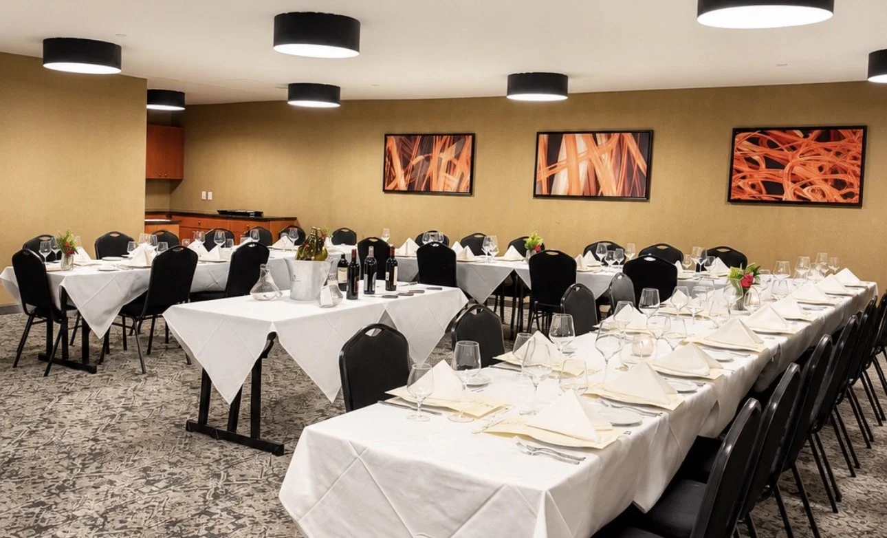 A formal dining setup in a banquet room with white tablecloths, wine glasses, and folded napkins on each plate. There are black chairs around the tables, and small floral centerpieces with red and green decorations. The room has beige walls, three abstract art pieces, and black ceiling lights.