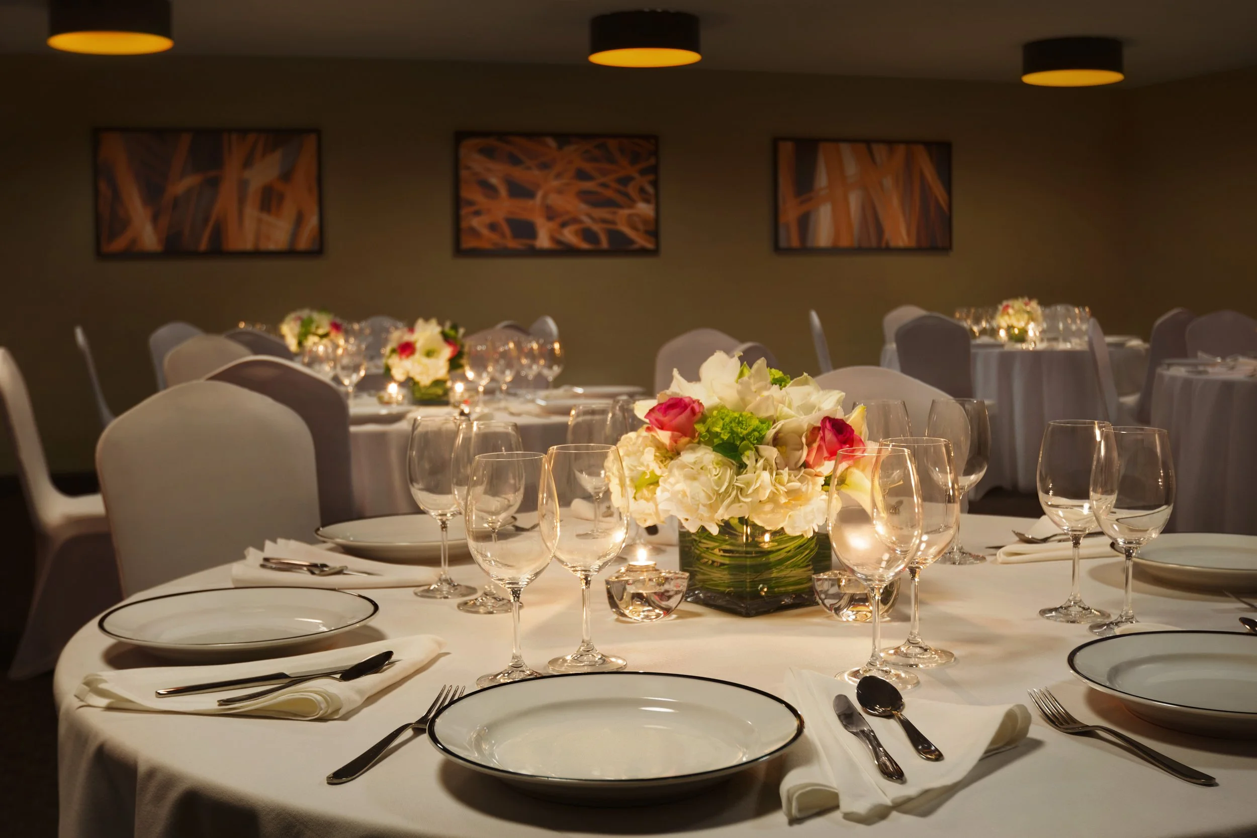 Elegant banquet table set with white tablecloth, floral centerpiece, multiple wine glasses, plates, and utensils in a dimly lit room.
