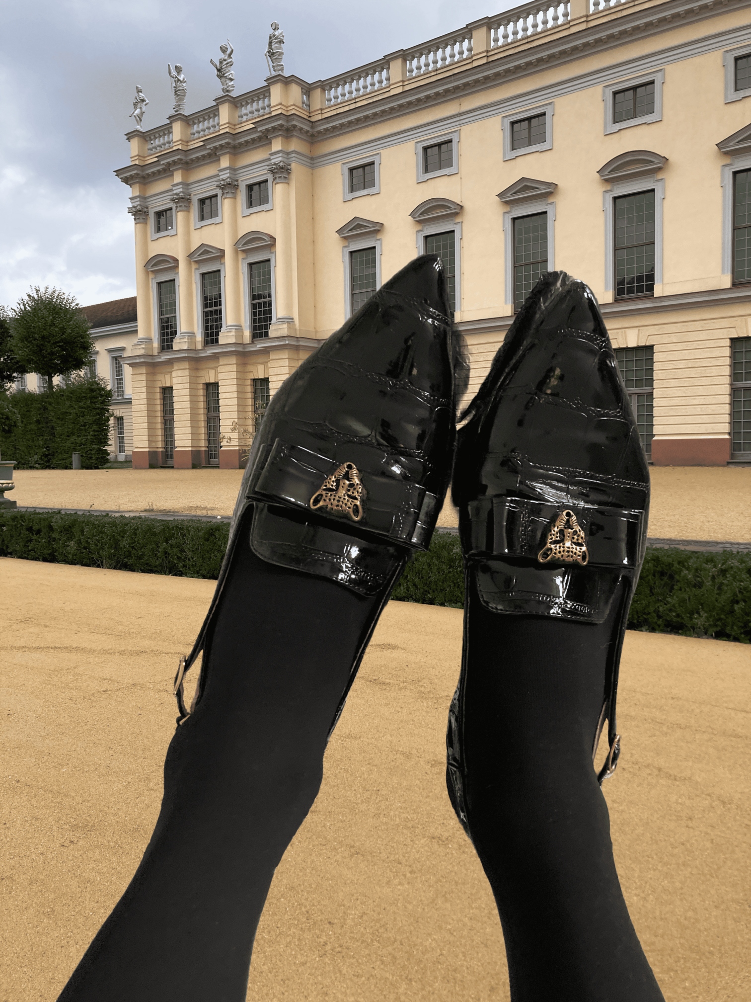 Ave Vanilla - Mademoiselle: An artistic composition of black patent leather loafers held up against the architectural backdrop of a historic palace facade[cite: 42].