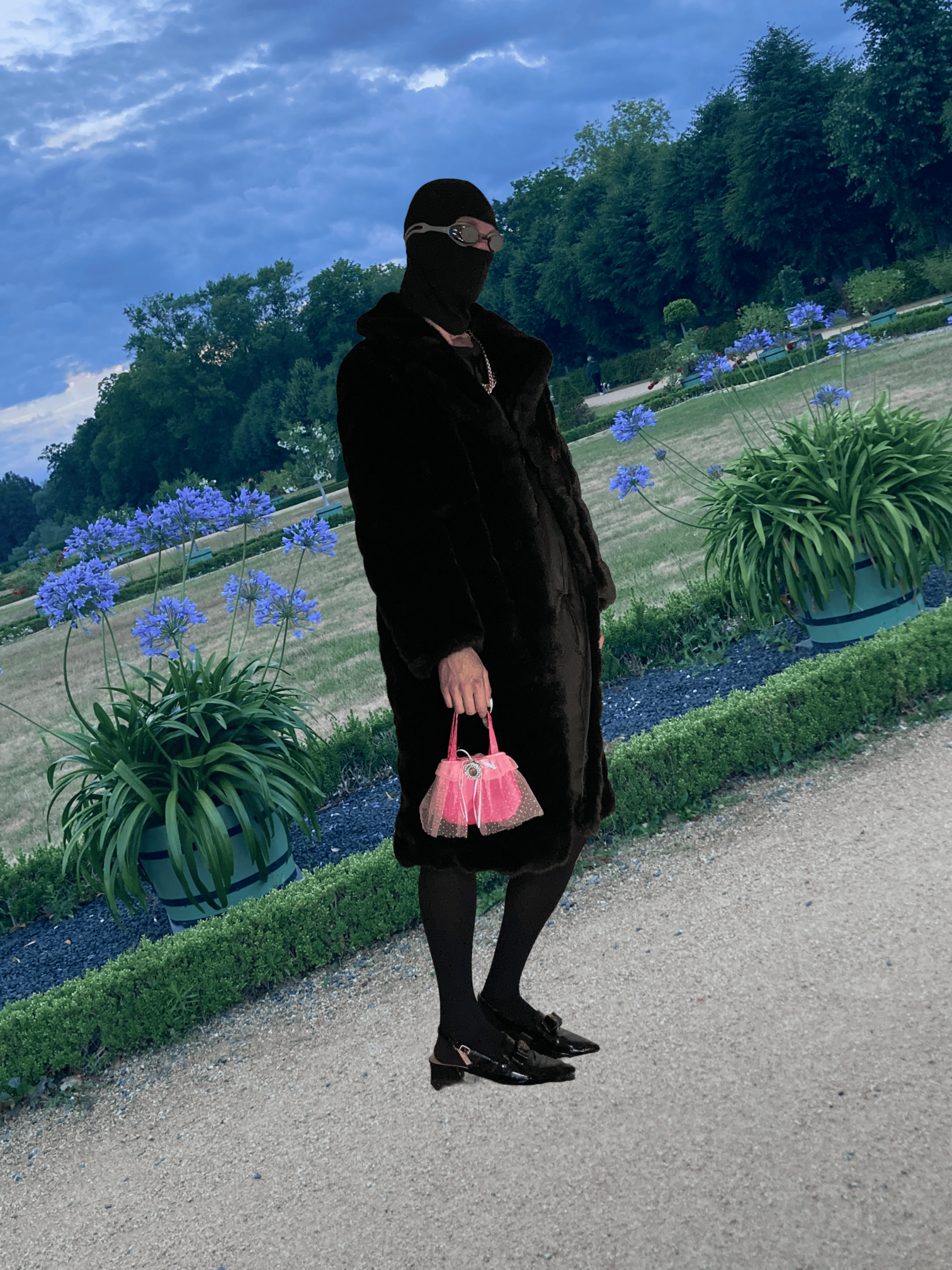 Ave Vanilla - Mademoiselle: A figure in a dark coat and balaclava holding a pink handbag, posed within the classical gardens of a grand palace[cite: 41].