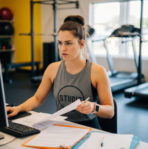 A fitness studio owner looking stressed at a front desk, trying to reconcile membership payments on a computer with a class in session in the background.