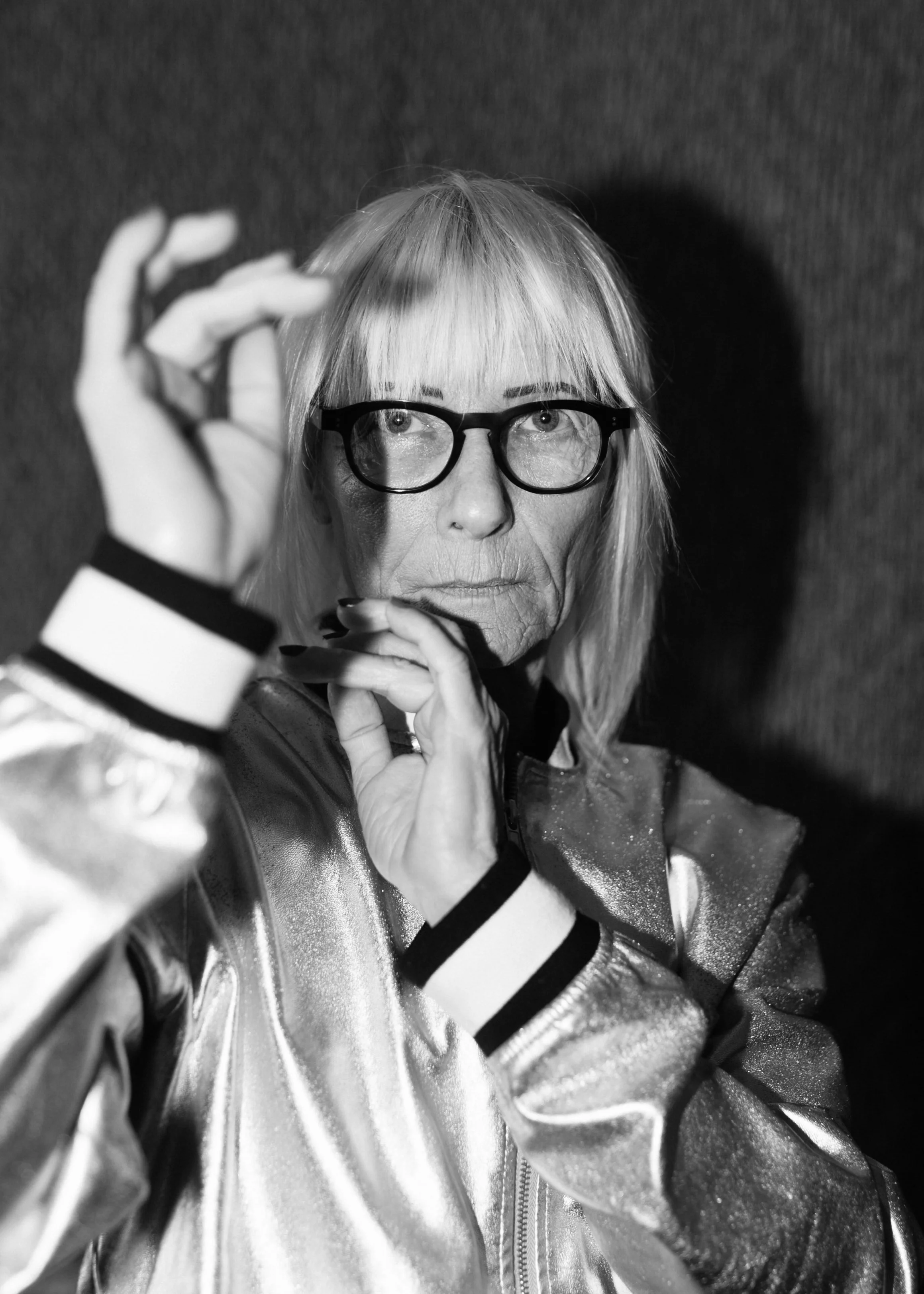 Black and white portrait of a person with glasses wearing a shiny jacket, holding up their hands contemplatively.