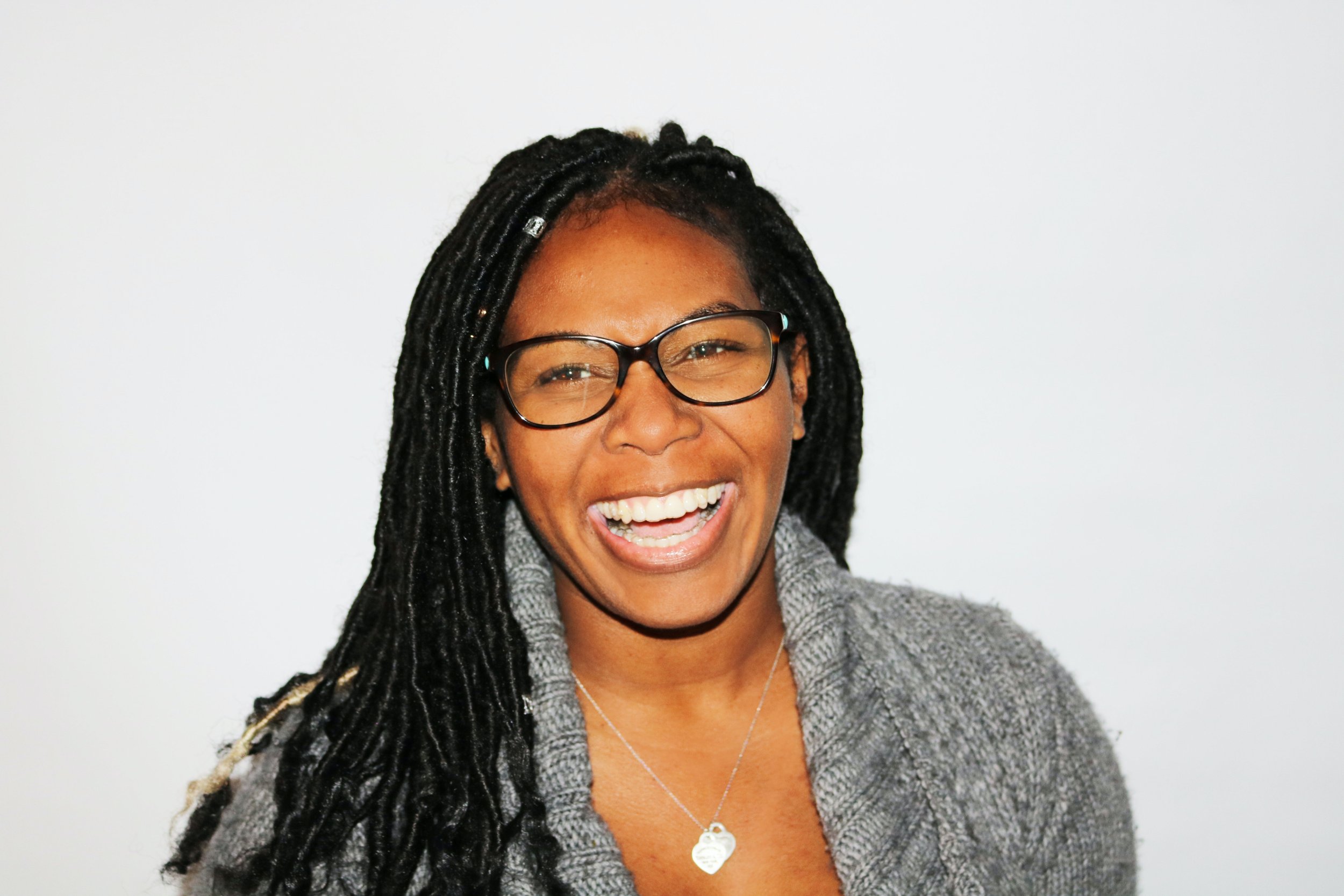Woman with glasses and dreadlocks smiling, wearing a gray sweater.