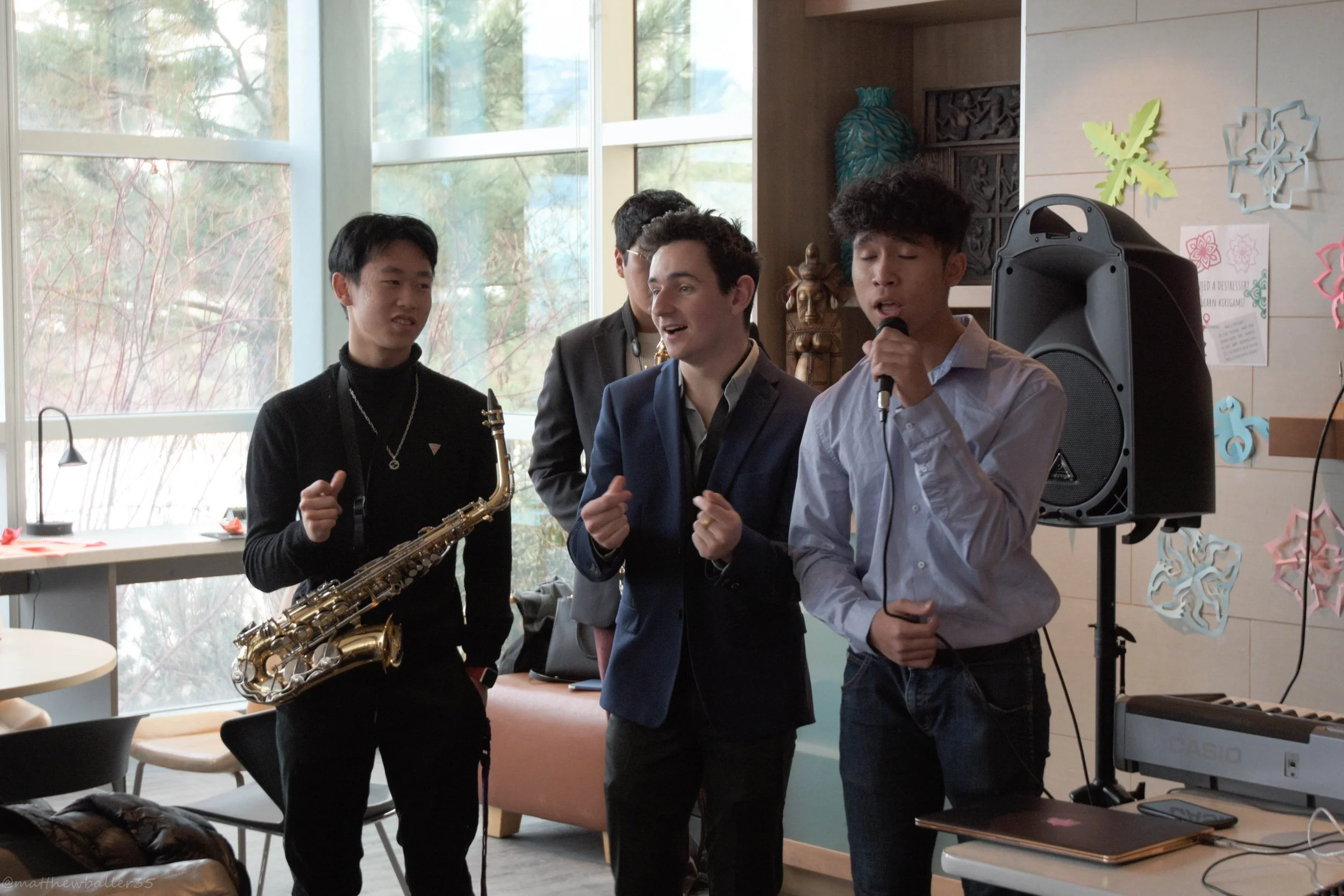 Four young men singing and playing music in a room with large windows, musical instruments, and colorful wall decorations.
