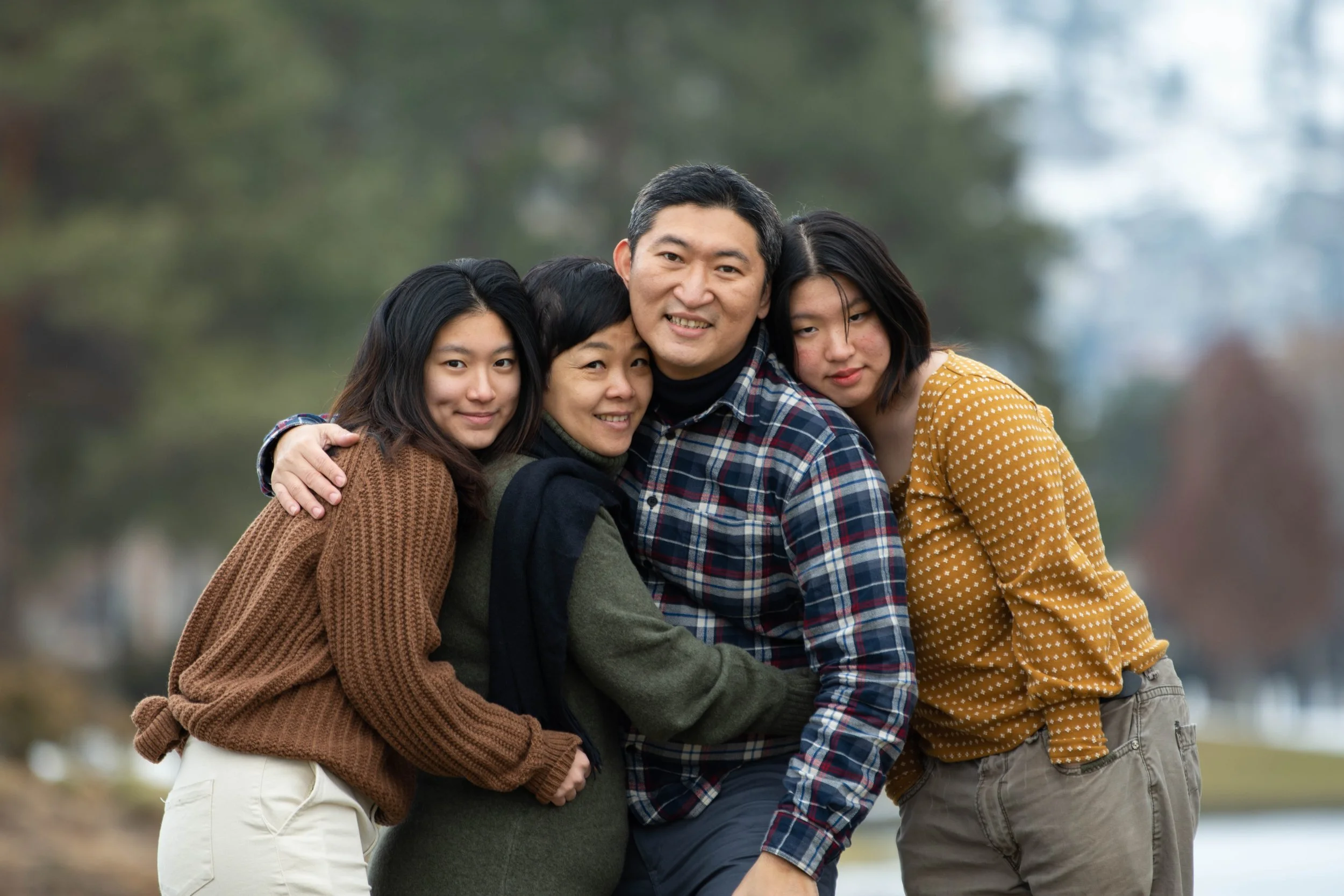 A family of four hugging outdoors during fall, with blurred trees in the background.