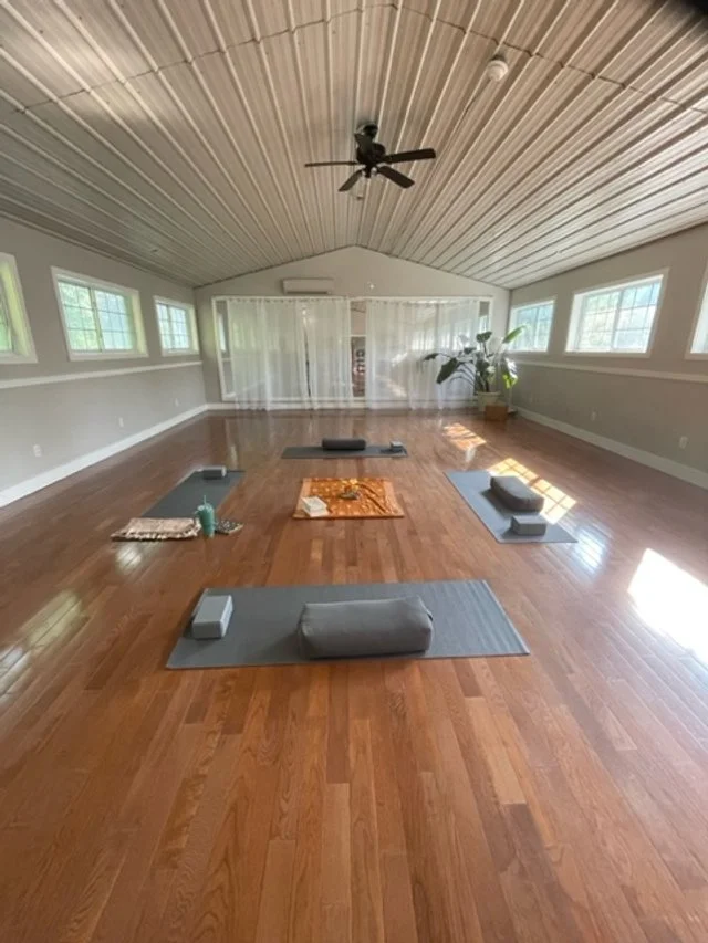 A spacious room with hardwood floors, a high wood-paneled ceiling, and multiple windows allowing natural light. The room is set up for yoga or meditation with mats, bolsters, and blocks arranged on the floor.
