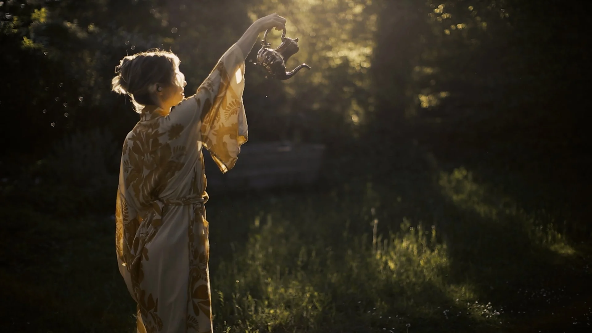 Frau mit einem goldfarbenen Kimono, die Wasser aus einer Teekanne auf einem sonnigen Waldweg gießt.
