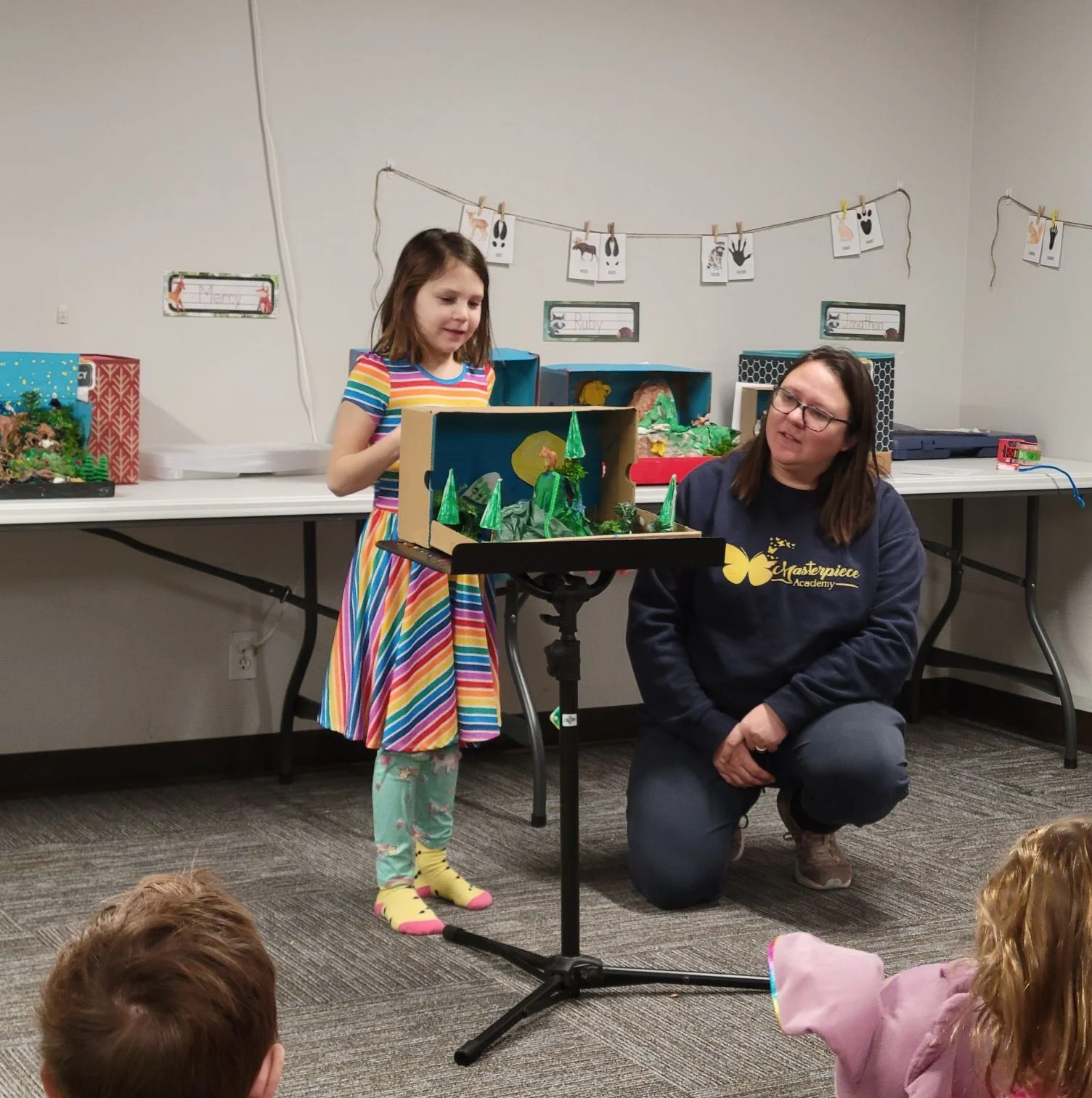 A young girl in a colorful striped dress and knee socks stands next to a woman kneeling beside a table, showing a diorama with trees and a moon. The scene is set in a classroom or library with other dioramas on the table and children watching in the