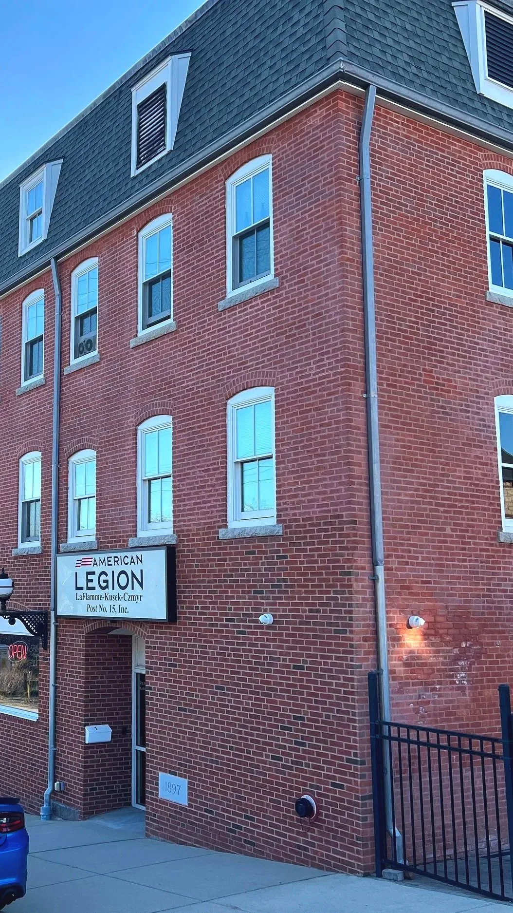 A multi-story brick building with several white-framed windows and a black sign that reads "American Legion LaFlamme-Kusek-Czmur Post No. 15, Inc." The building has a dark gray shingled roof with a vent, a sidewalk in front, and a gated entrance to the right. An